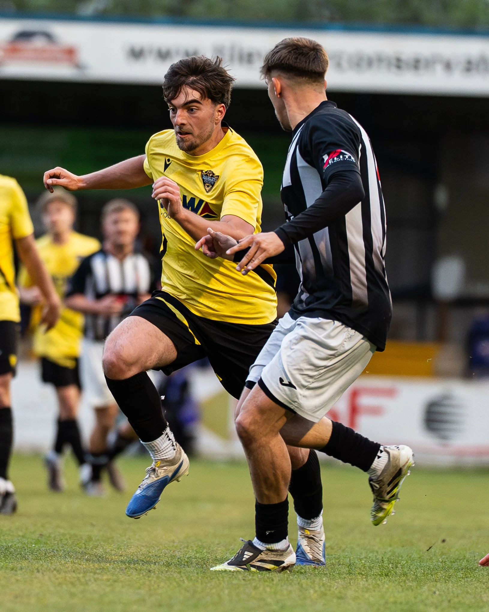Two soccer players running on the field during a game, with the player in yellow focused and the player in black and white attempting to keep up, with blurred background of other players and spectators.