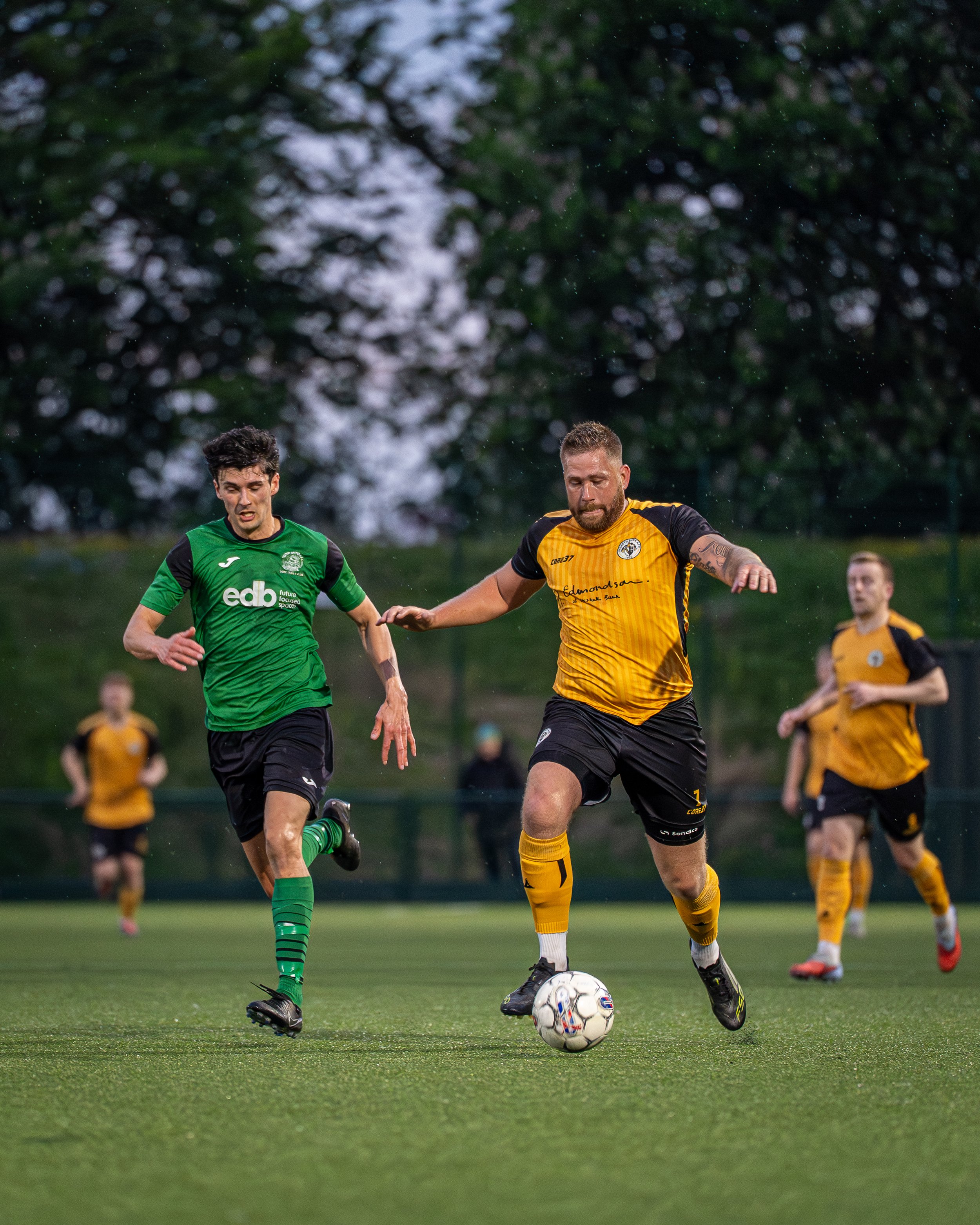 Two soccer players, one in a green uniform and one in a yellow and black uniform, competing for the ball on a grassy field with a blurred background of other players and trees.