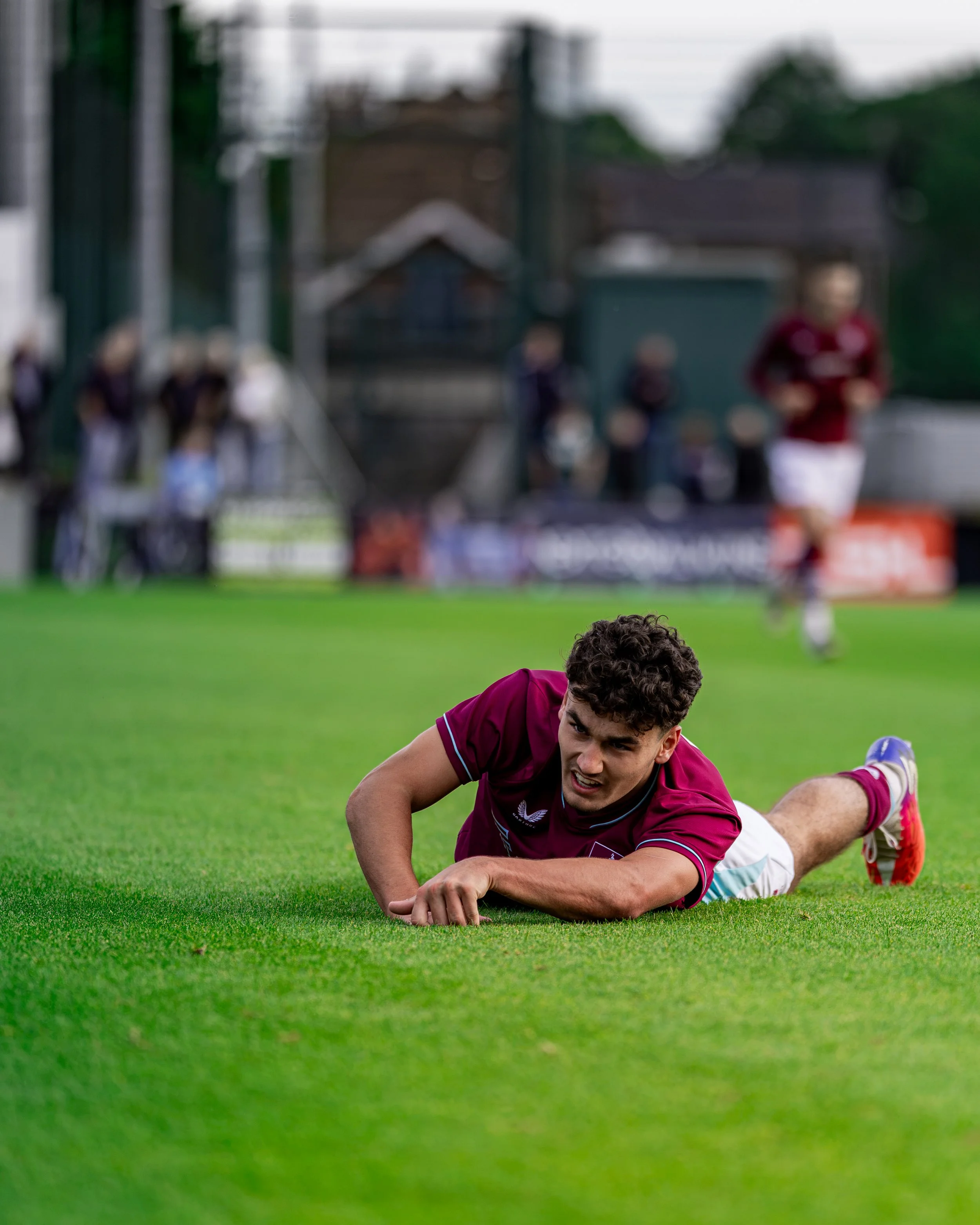 A soccer player in maroon uniform falling on the grass during a game, with other players and a blurred spectator stand in the background.