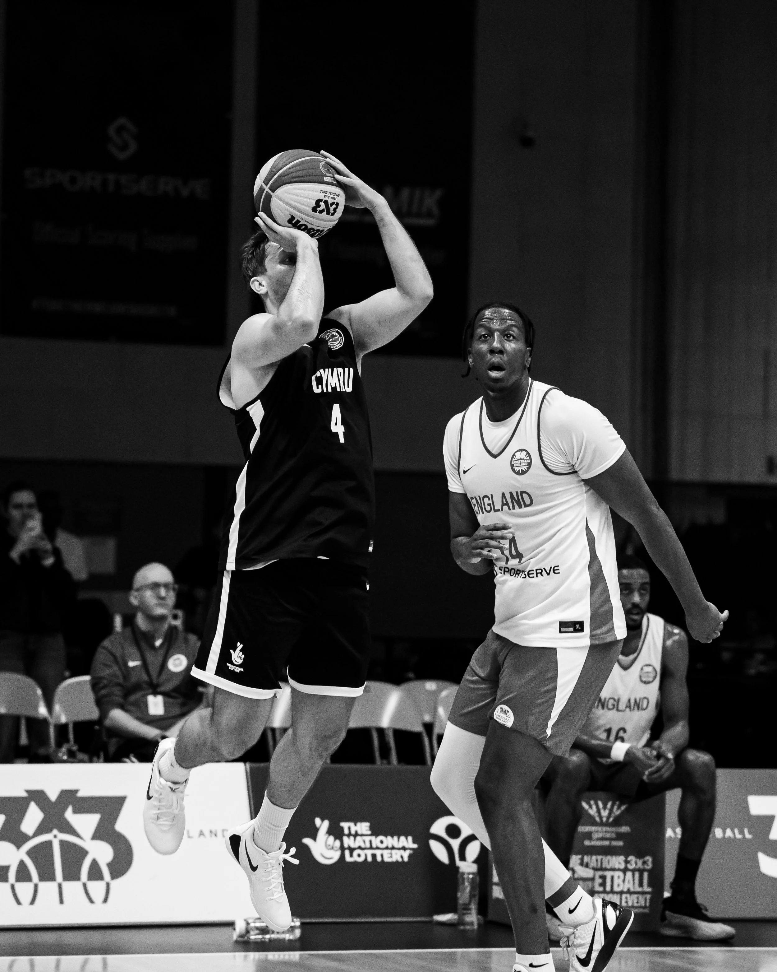 Black and white photo of a male basketball player in the middle of a shot during a game, with an opponent in front and spectators in the background.