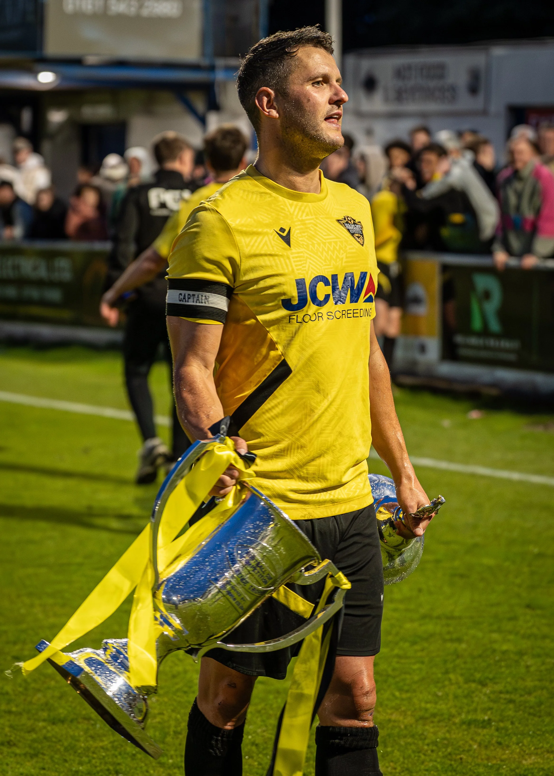 A male soccer player in a yellow jersey with a captain's armband, holding a trophy, on a grass field with a crowd in the background.
