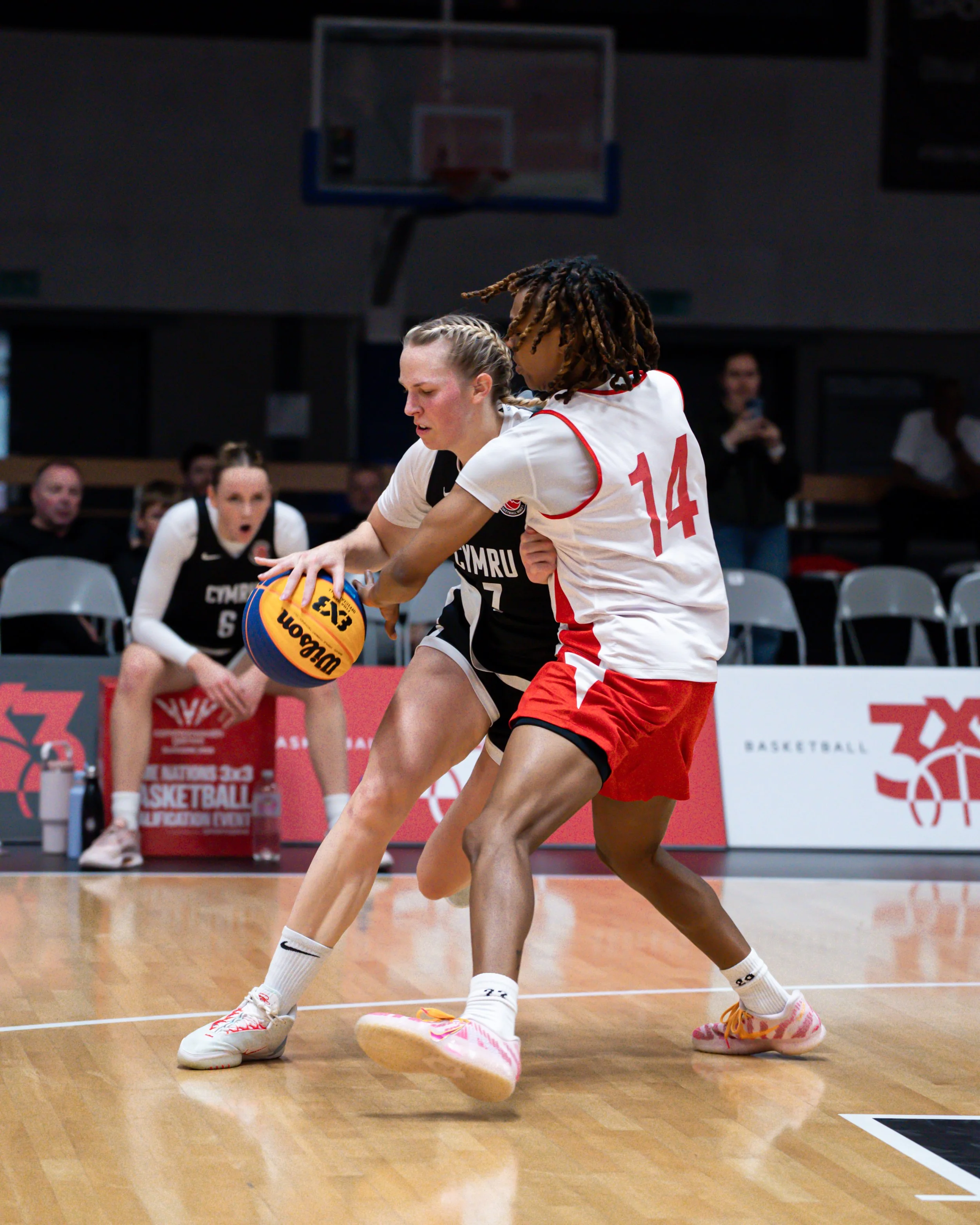 Two female basketball players, one in black and one in white and red. The player in black is holding a Wilson basketball, and both are fighting for possession on the court.