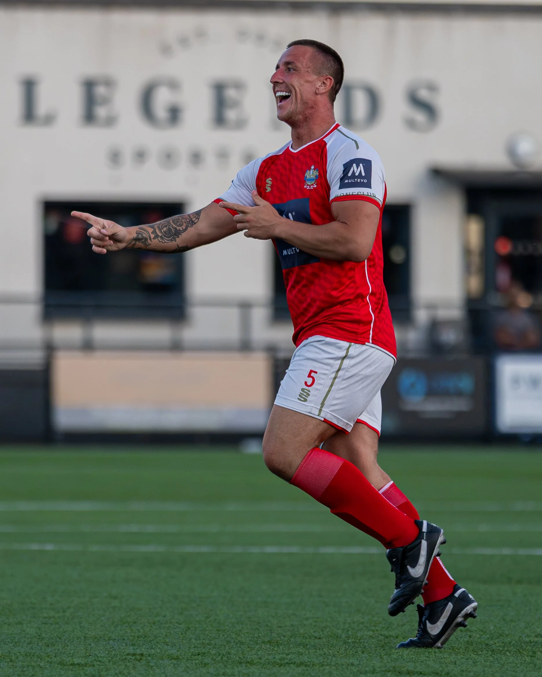 A soccer player wearing a red and white uniform with the number 5, celebrating with a big smile, pointing with both hands during a match on a green field.