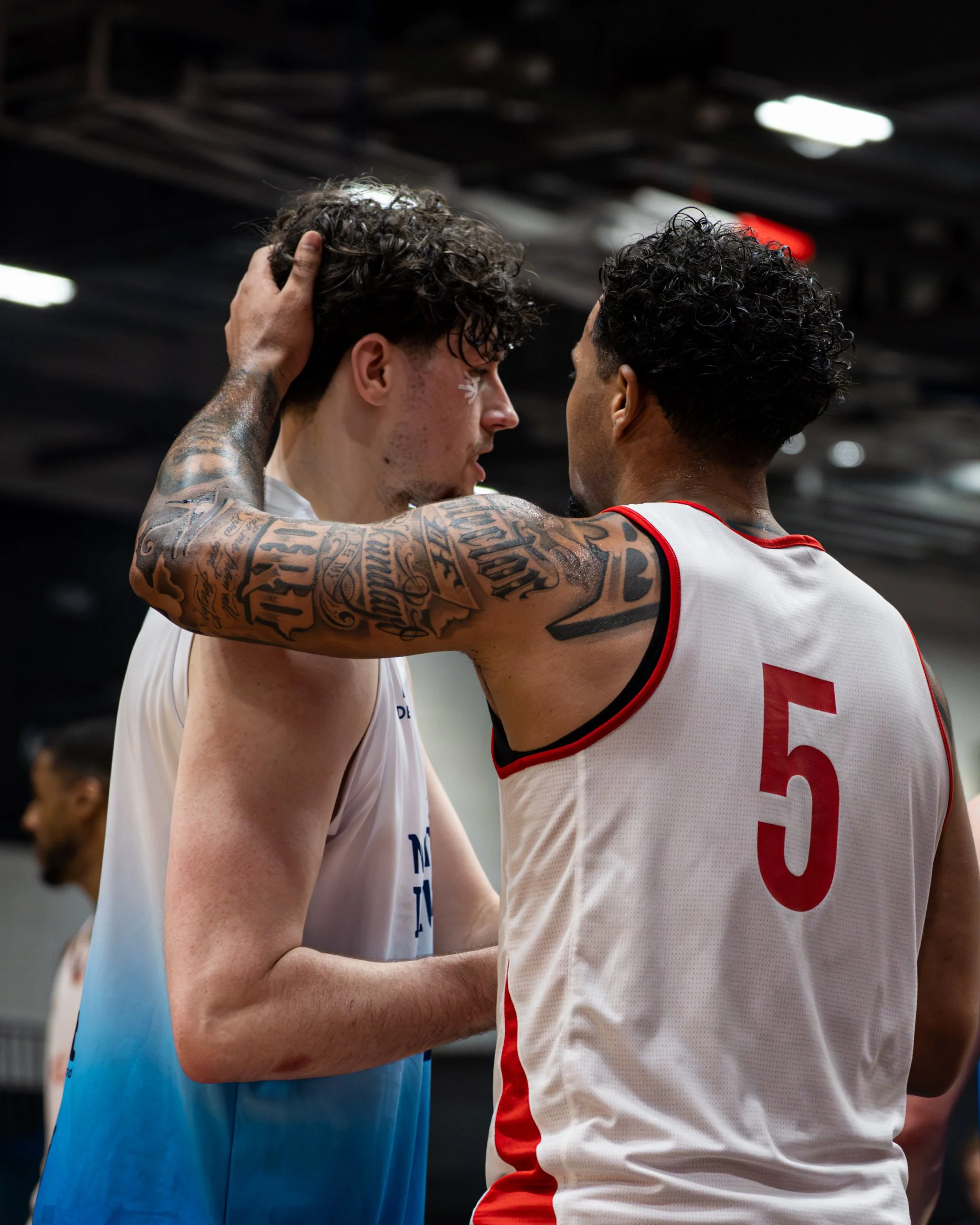 Two basketball players, one in a white jersey with red trim and the other in a white and blue jersey, are facing each other closely, with one player touching the other's head in a gesture of respect or comfort.