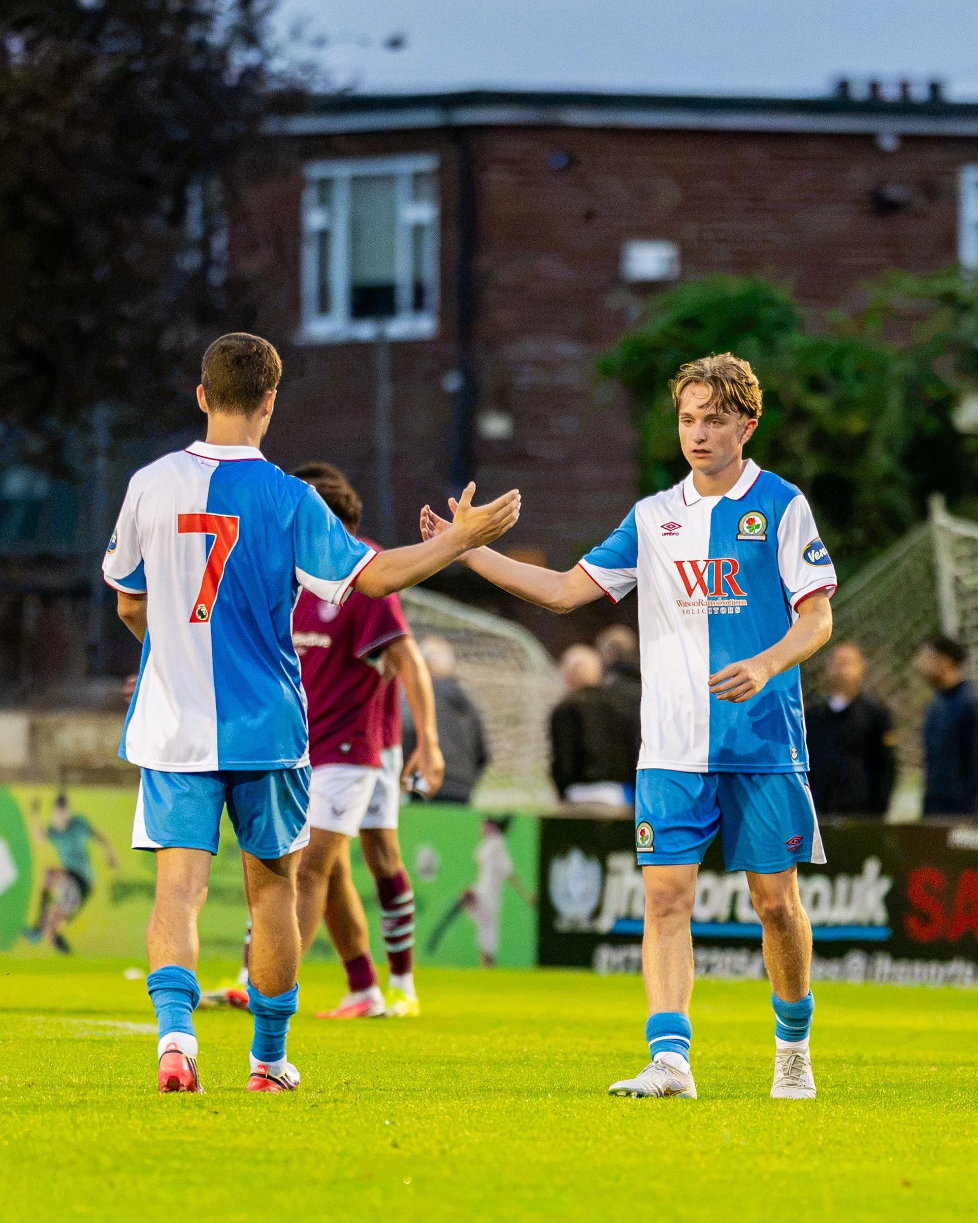 Soccer players in blue and white uniforms exchanging high-fives on a green field, with a brick building and spectators in the background.