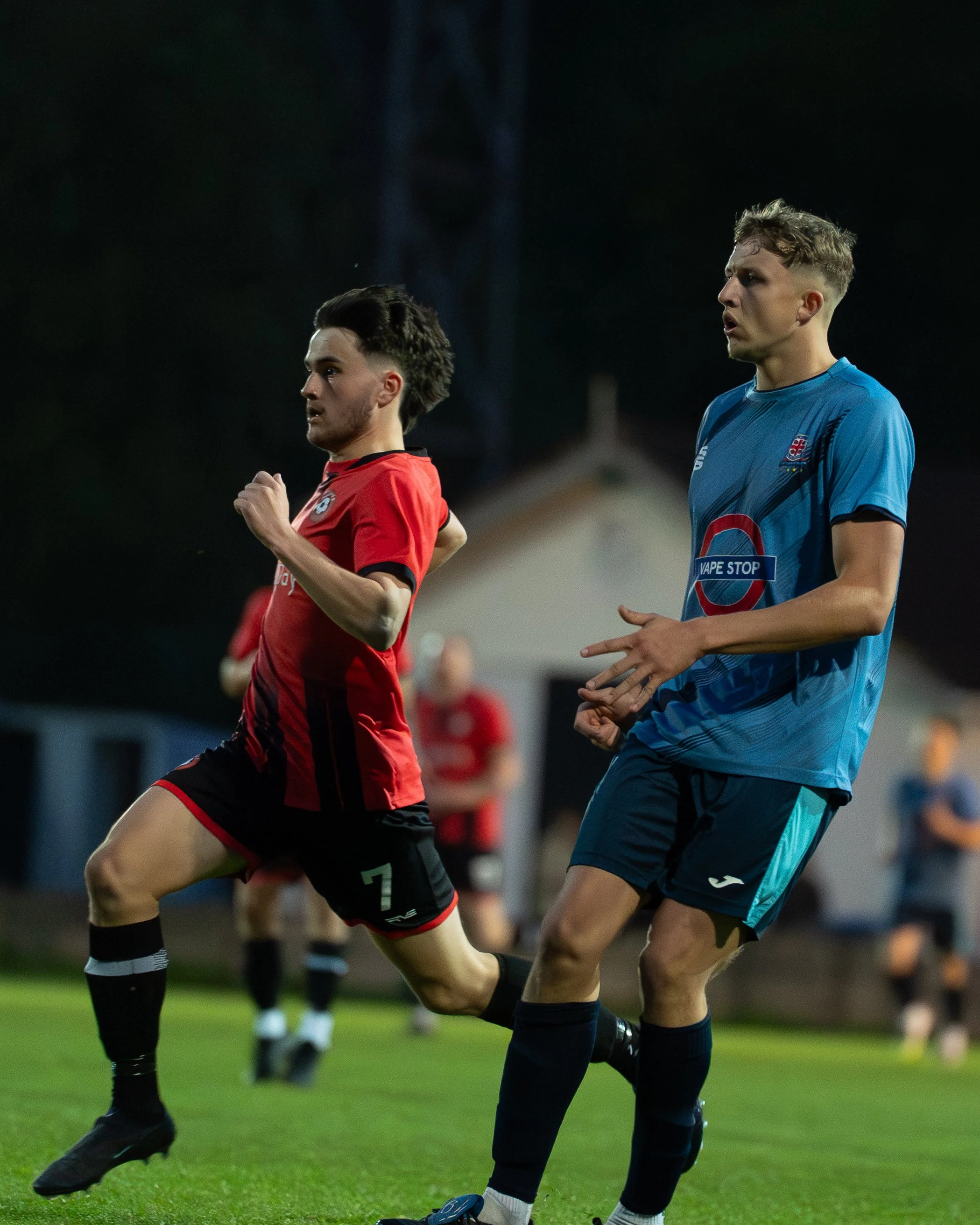 Two male soccer players running on a field during a game at night, with a dark sky and fields in the background. One player wears a red and black uniform, and the other wears a blue uniform with a logo.
