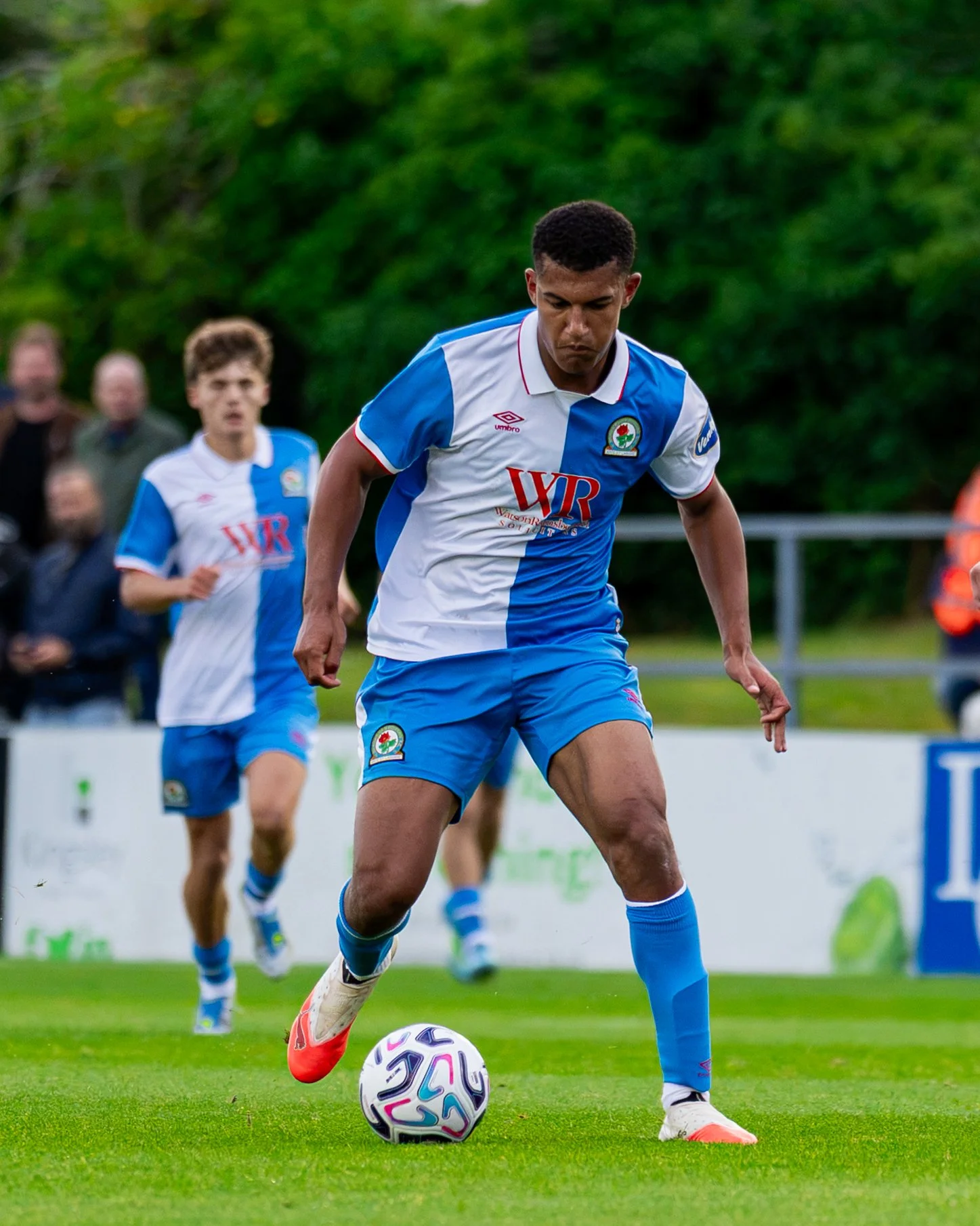 Soccer player in a blue and white jersey kicking a soccer ball on a field with spectators in the background.