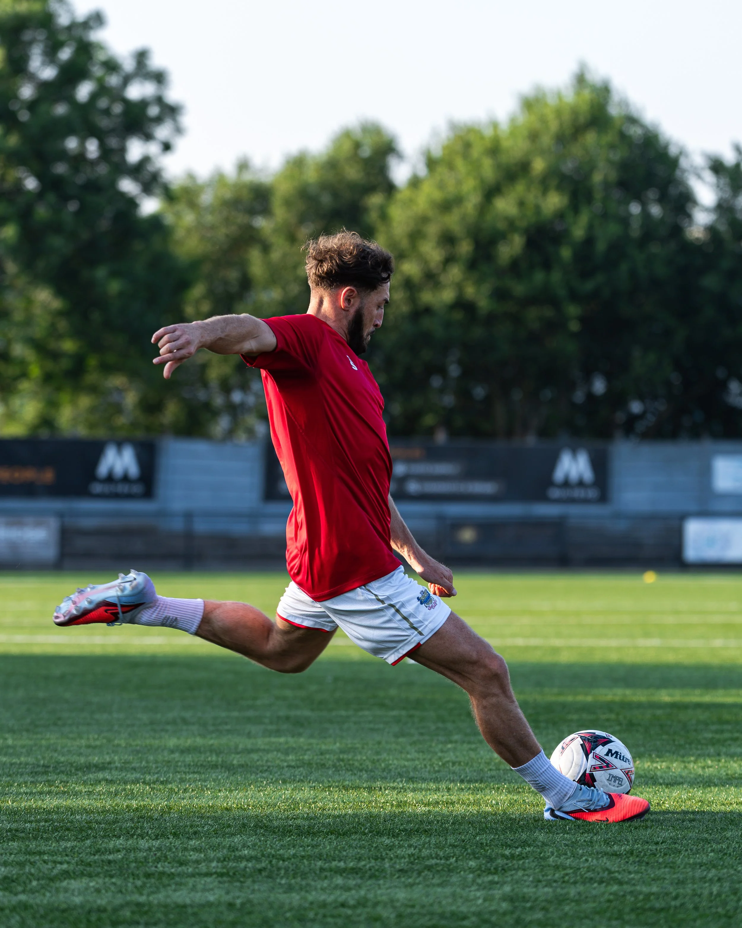 Male soccer player in a red shirt and white shorts kicking a soccer ball on a green field with trees in the background.