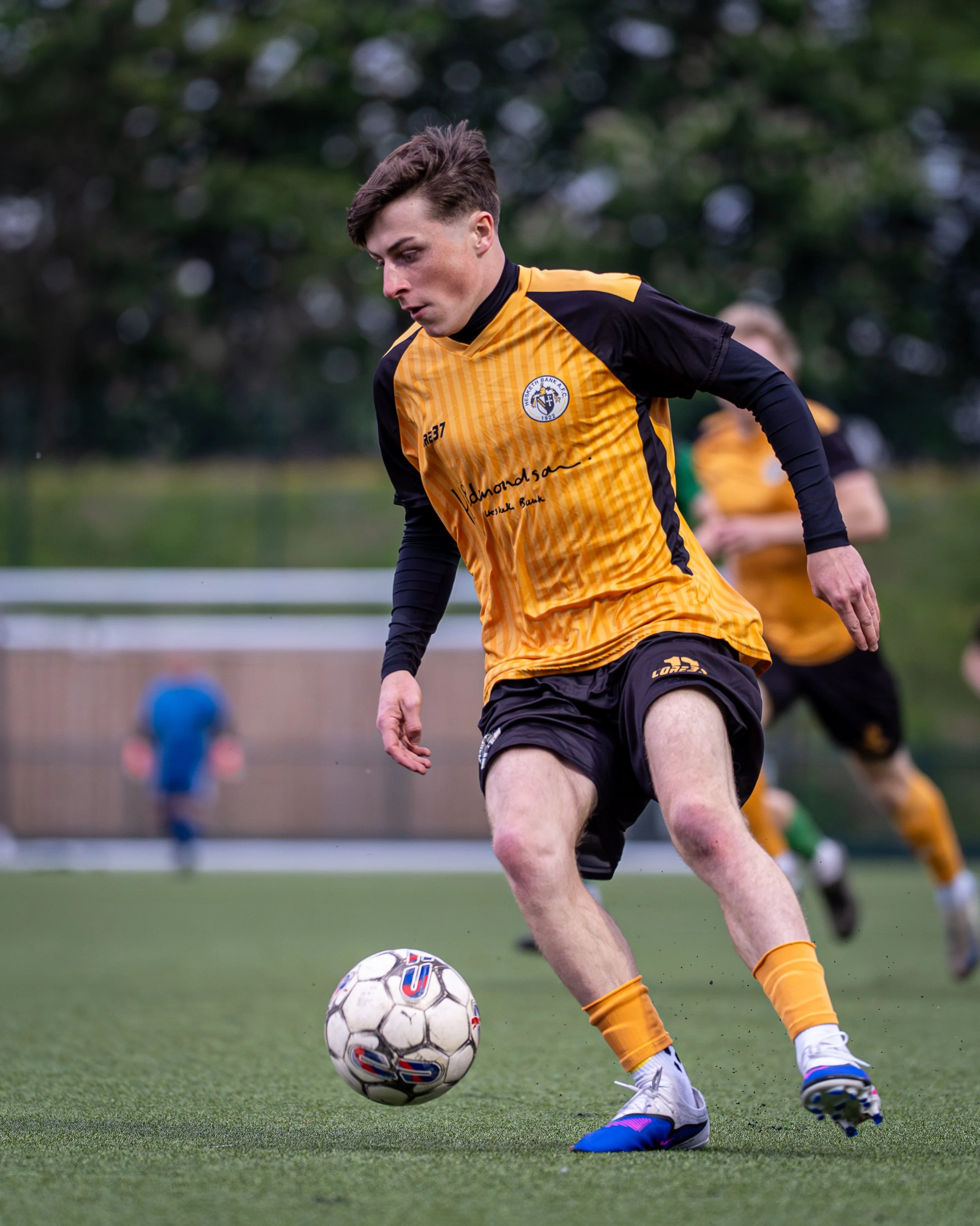 A young male soccer player in yellow and black uniform dribbling a soccer ball on a green field with trees in the background.