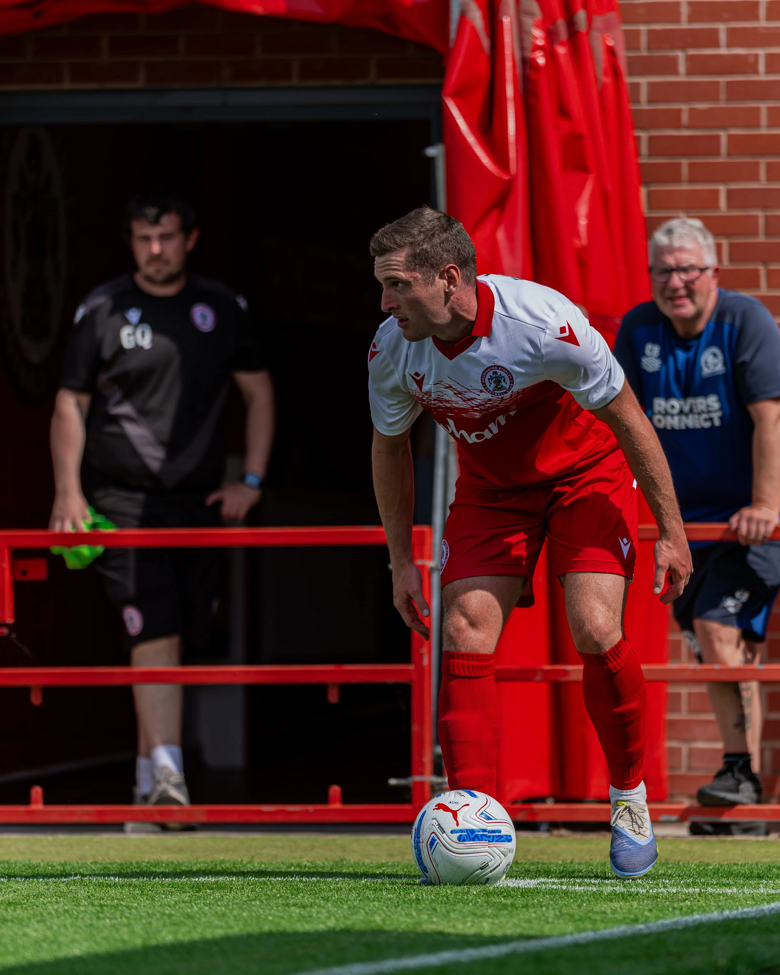 A soccer player in a red and white uniform is about to kick a soccer ball on a field. Behind him, two men are seated on a bench, watching. The background includes a brick wall and red structures.