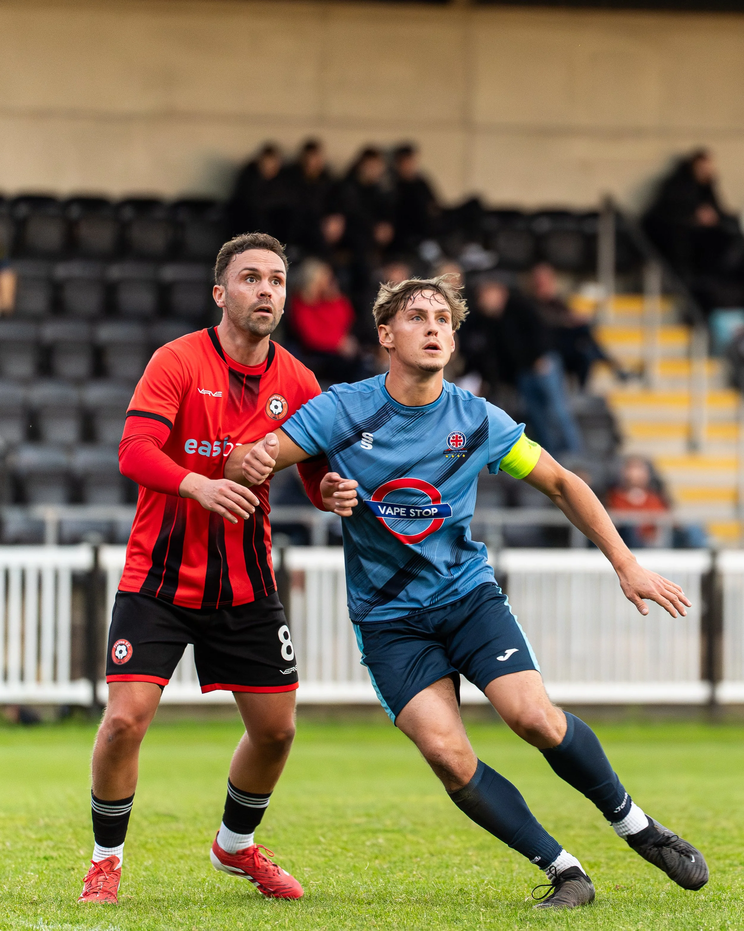 Two male soccer players in action on the field, one wearing a red jersey and the other in a blue jersey, competing for the ball during a match.