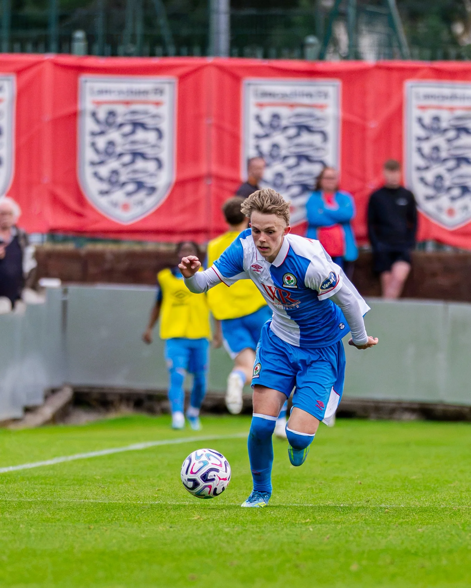 A young male soccer player with blonde hair, wearing a white and blue kit, is running on a grass field with a soccer ball at his feet. In the background, there are spectators and banners with a lion emblem.
