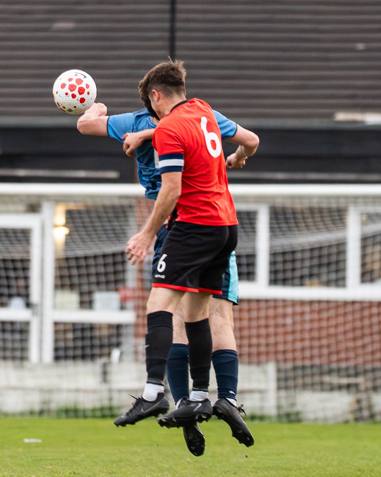Two soccer players jumping to head the ball during a match, with one wearing a red jersey with the number 6 and the other in a blue jersey, on a green field with goal targets in the background.