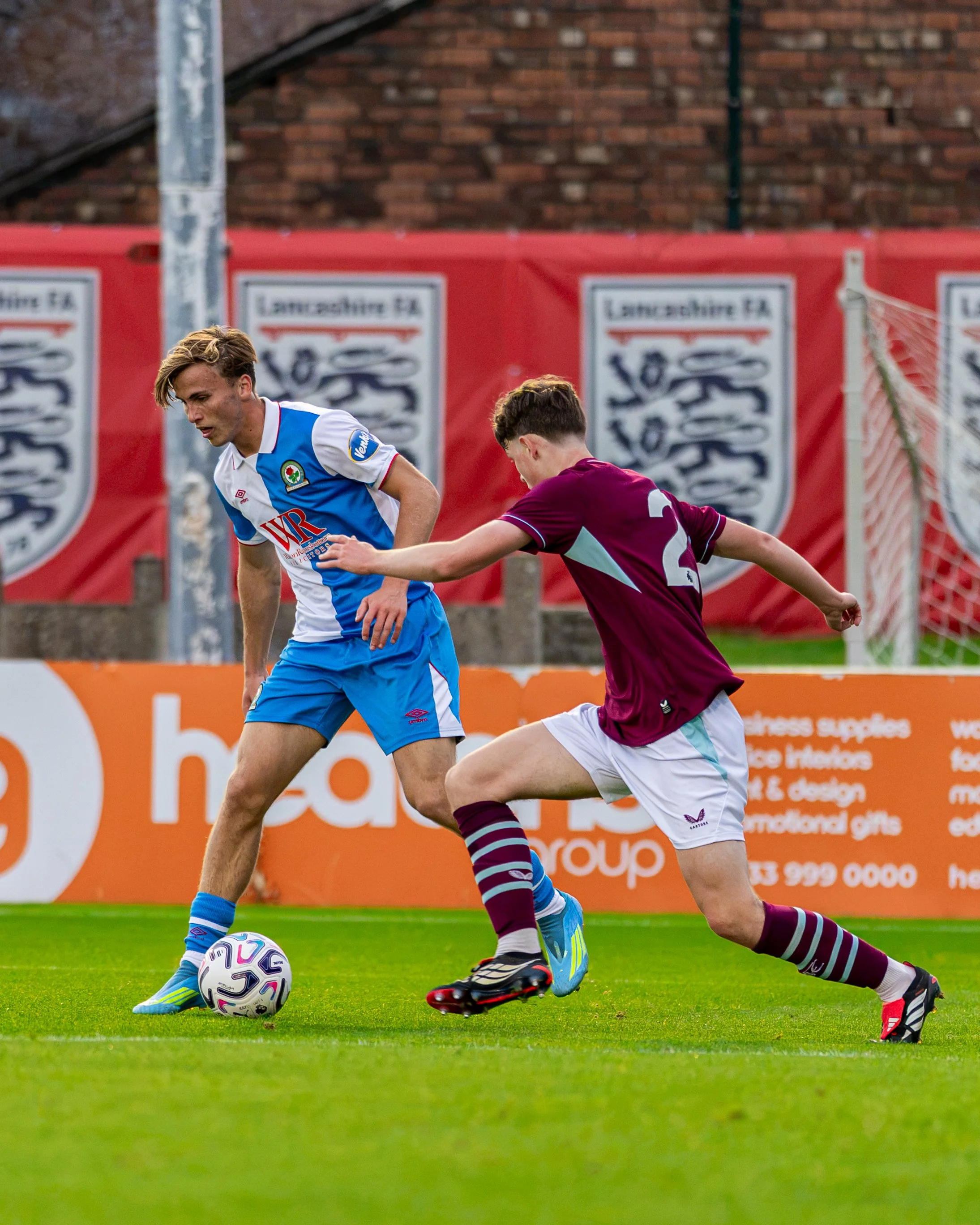 Two young soccer players competing for ball on field. One player wearing blue and white uniform, the other in maroon and teal uniform. Background features red advertising banners and brick building.