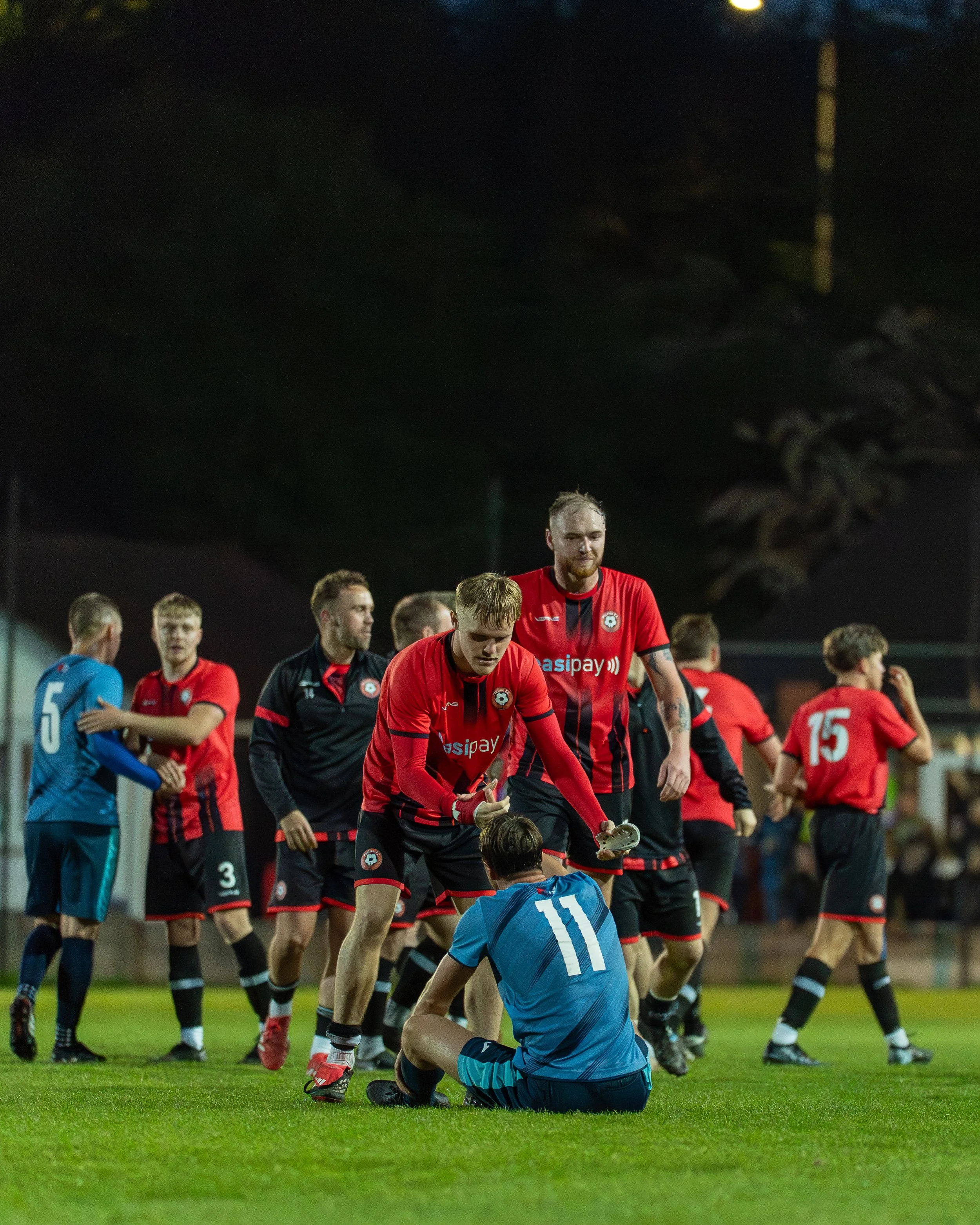 Soccer players on the field after a game, with some players in red jerseys and one in blue, under stadium lights at night.