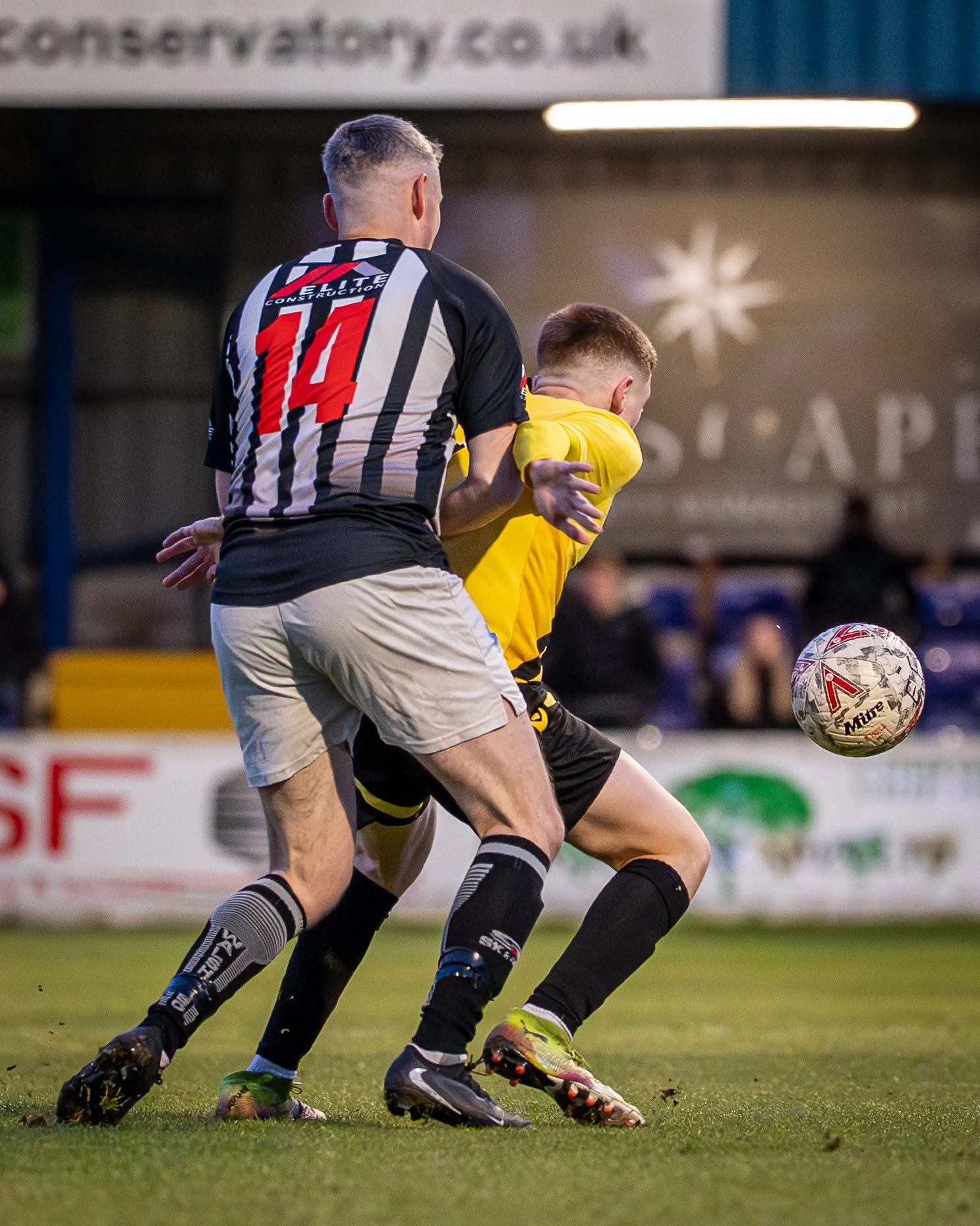 Two soccer players competing for the ball during a game, with one in a black and white striped jersey with red number 14 and the other in a yellow jersey. The scene is set on a grassy field with a blurred background and stadium advertisements.