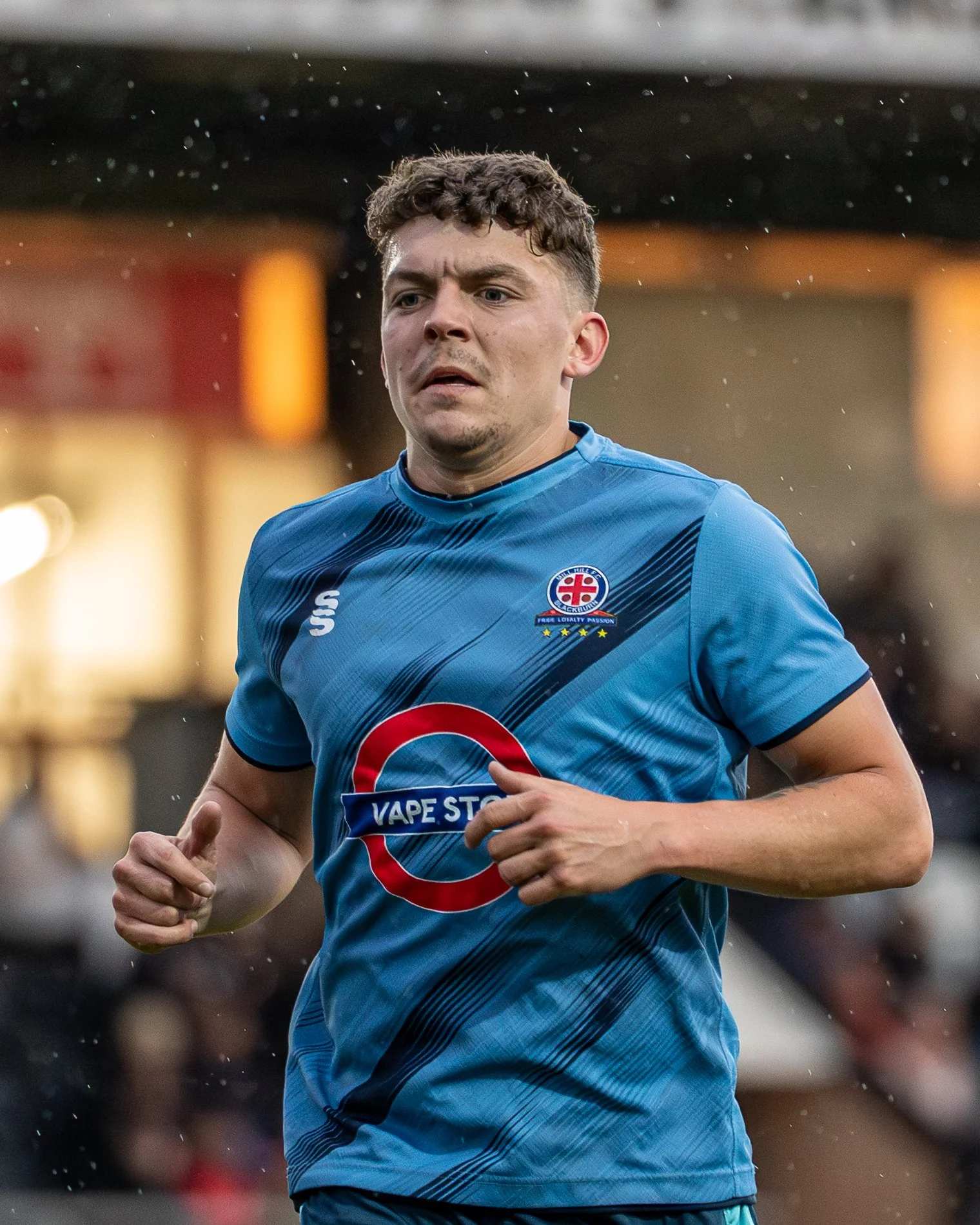 A man in a blue sports jersey running outdoors during rain, with blurred background and water droplets visible.