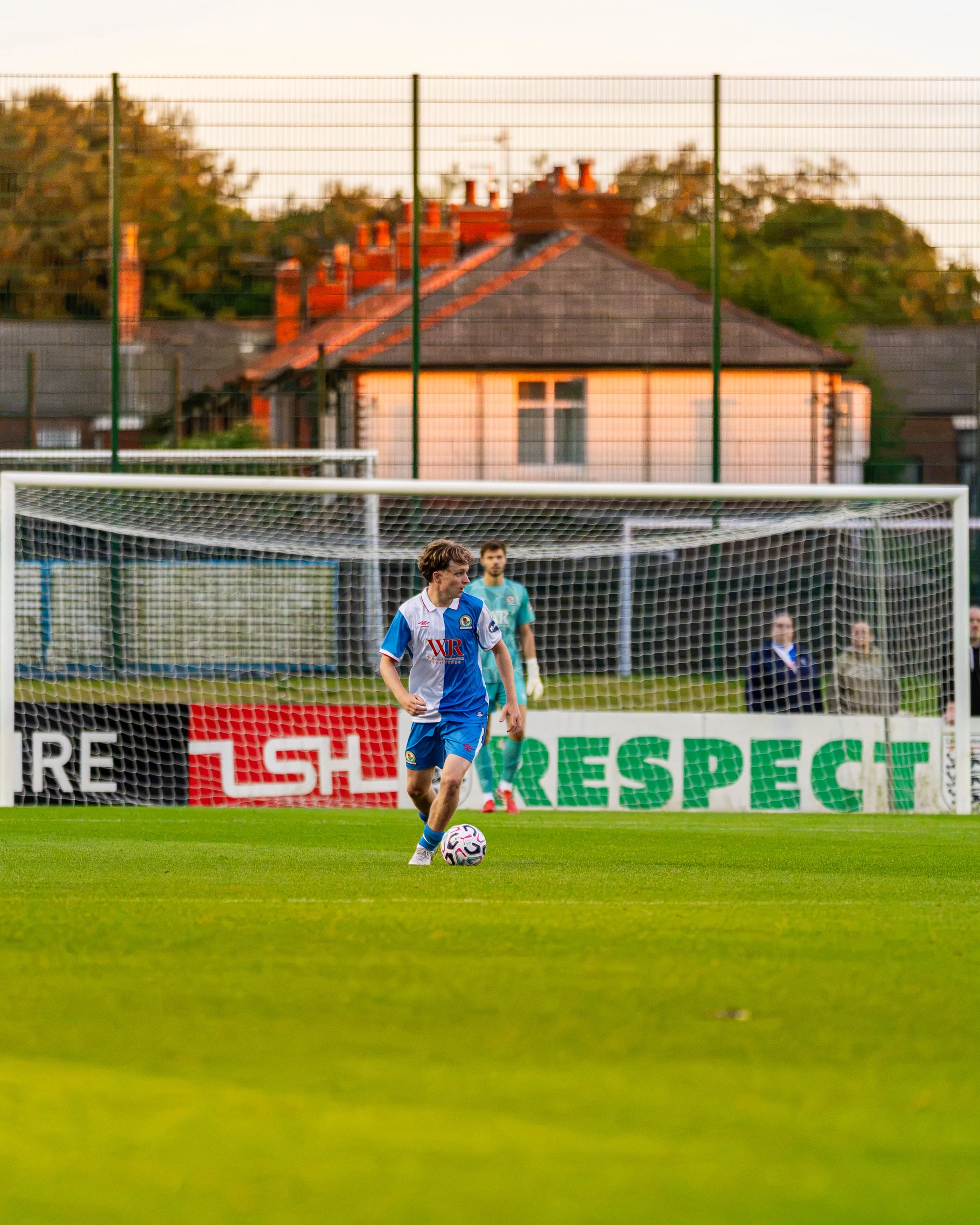 Soccer player in a blue and white jersey preparing to kick a ball on a soccer field during a late afternoon or early evening game, with a soccer goal in the foreground and residential houses and trees in the background.