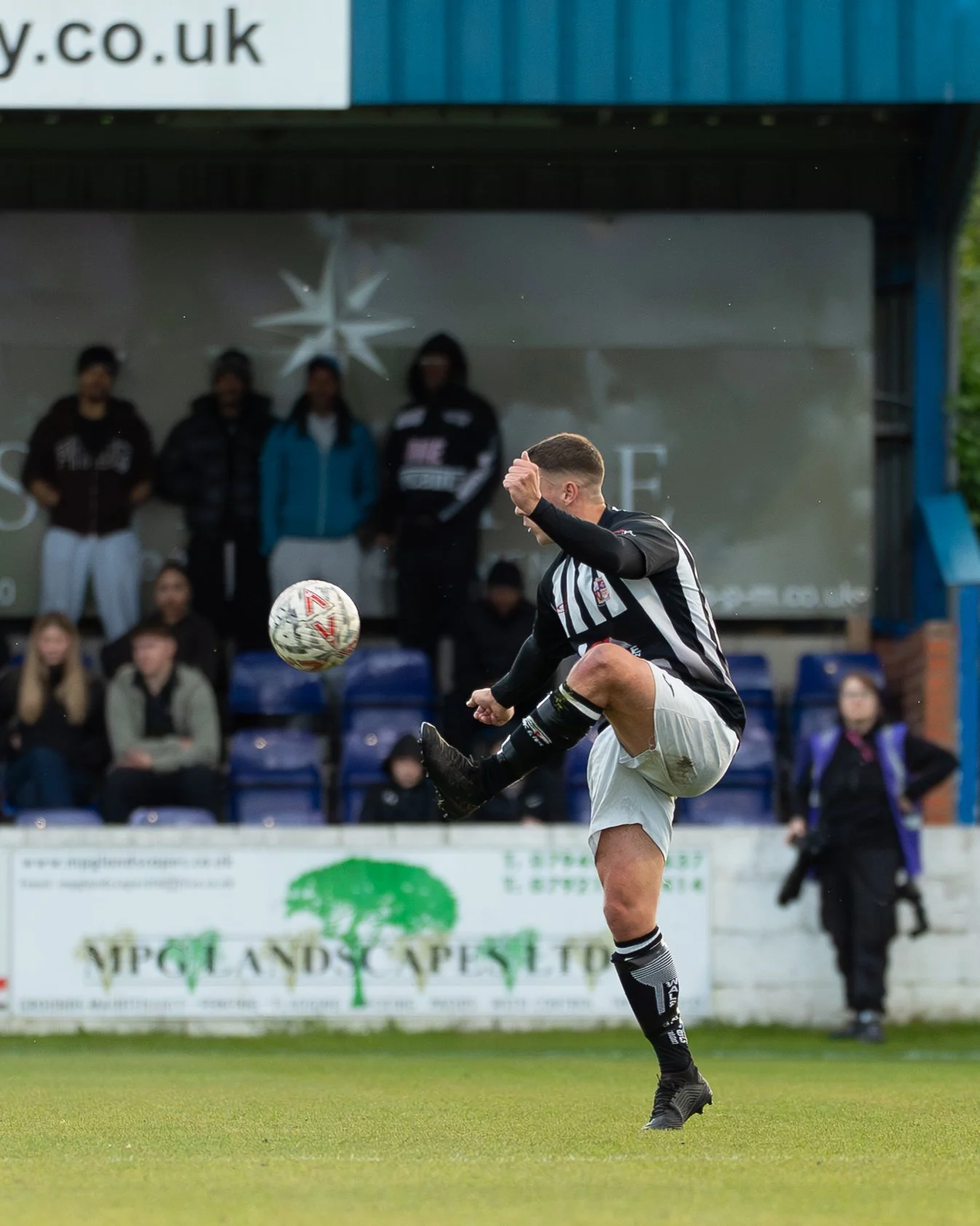 A soccer player in a black and white striped jersey kicks a soccer ball on the field, with spectators in the background.