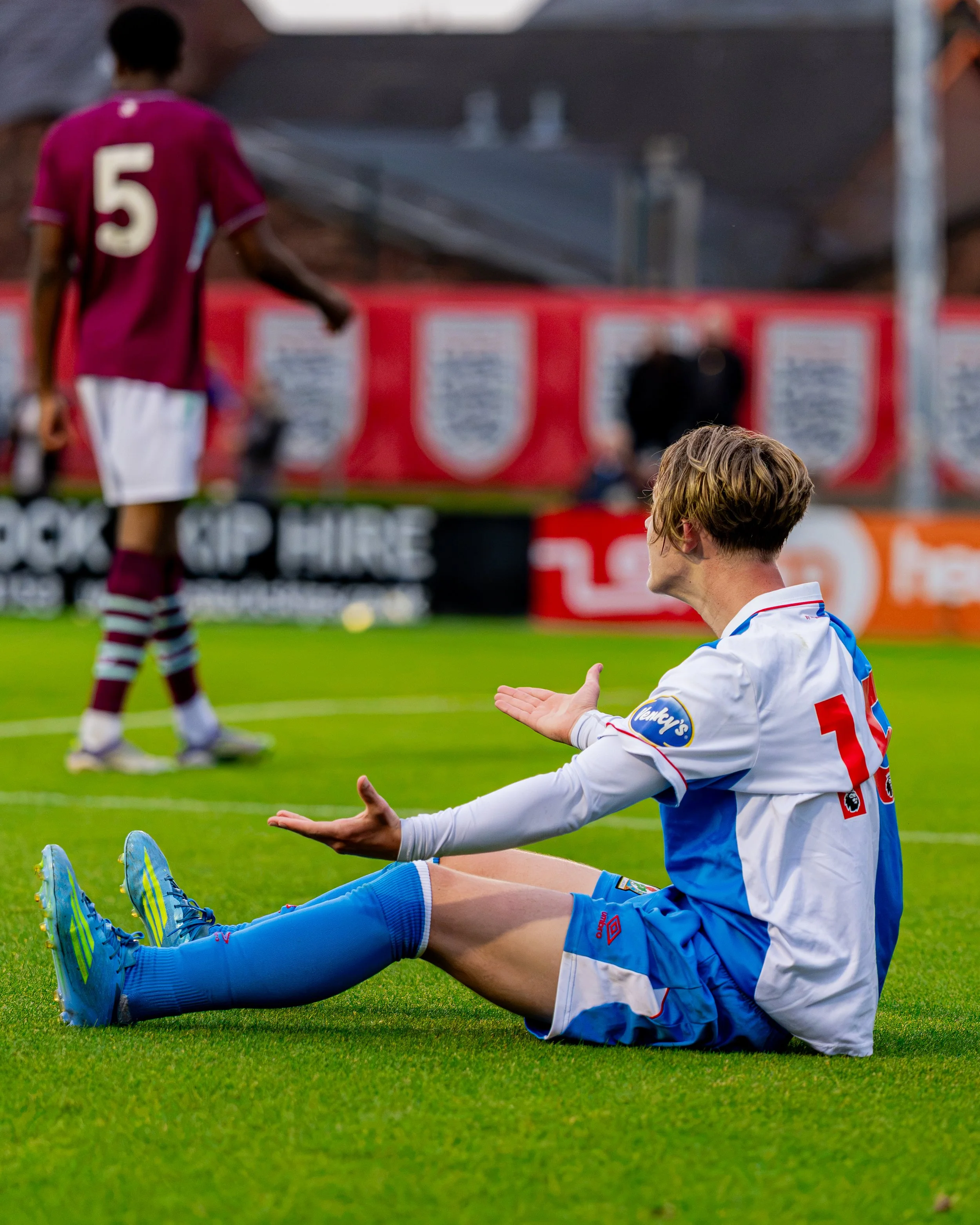 A soccer player in blue uniform sitting on the field with arms open, following a game, with another player in a maroon uniform walking in the background.