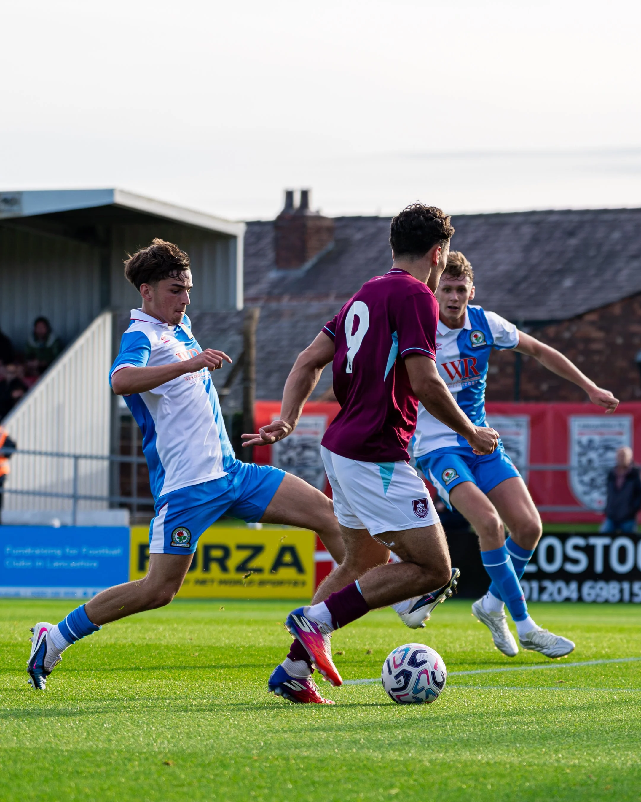 Three soccer players competing for the ball on a grassy field, with two wearing blue and white uniforms and one in maroon, during a match outdoors in the late afternoon.