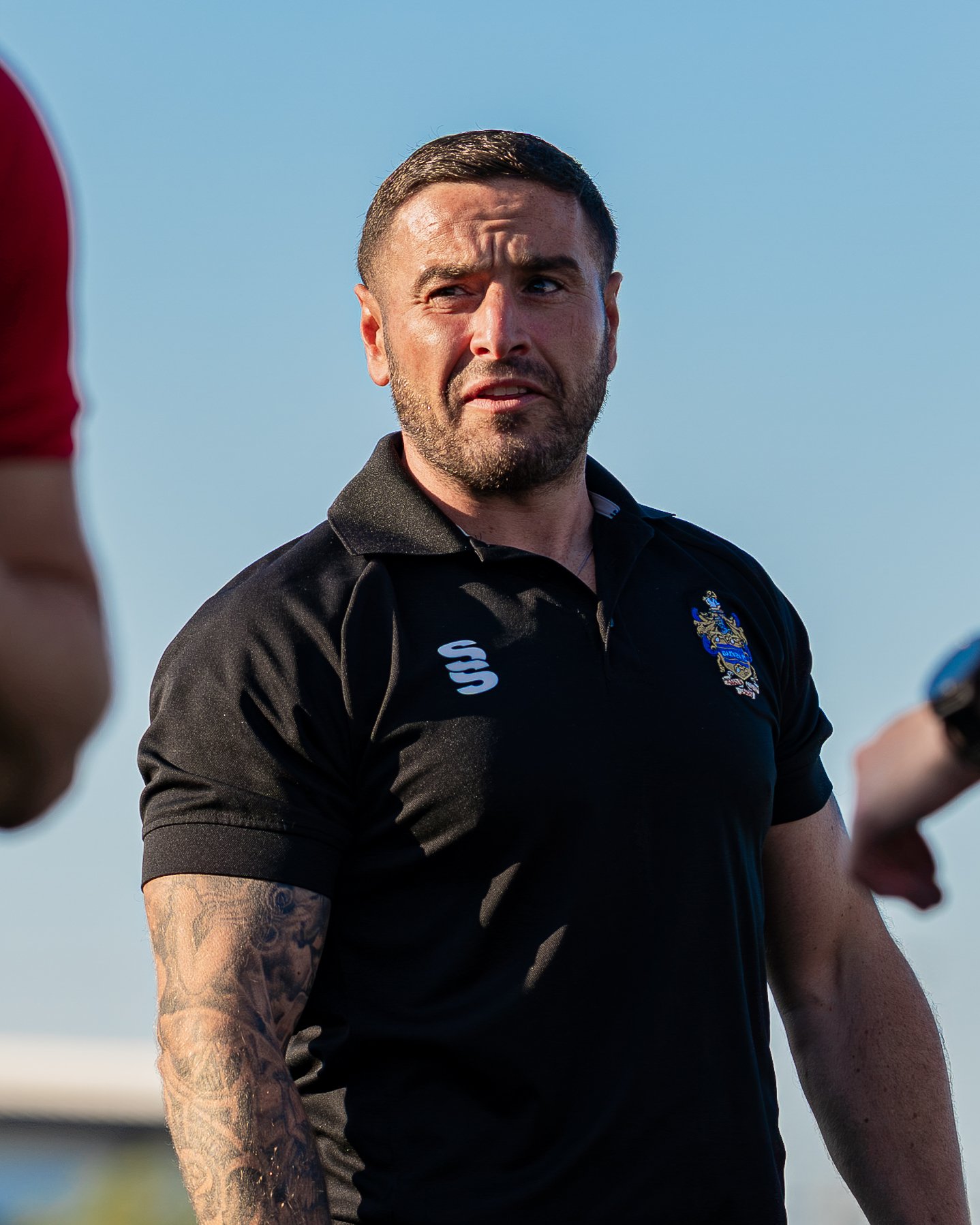 A man with tattoos on his left arm and a beard, wearing a black polo shirt with a crest, is outside under a clear blue sky. He appears to be speaking or reacting emotionally, with a serious expression, and is engaged in a conversation or activity.