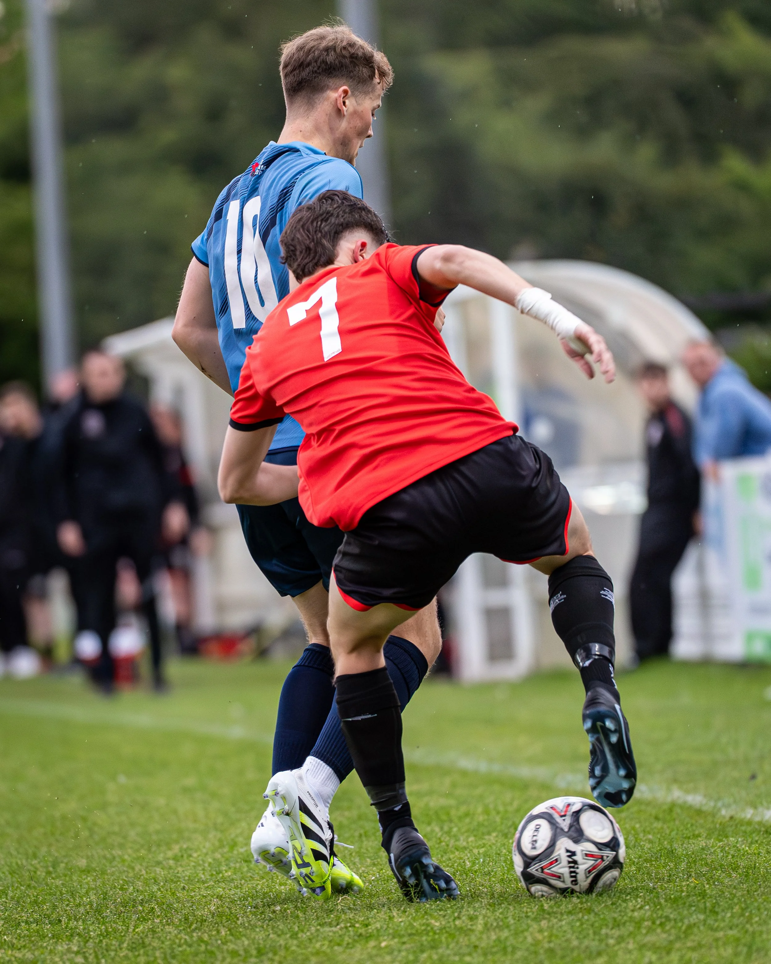 Two soccer players competing for the ball on a grassy field, with spectators and trees in the background.