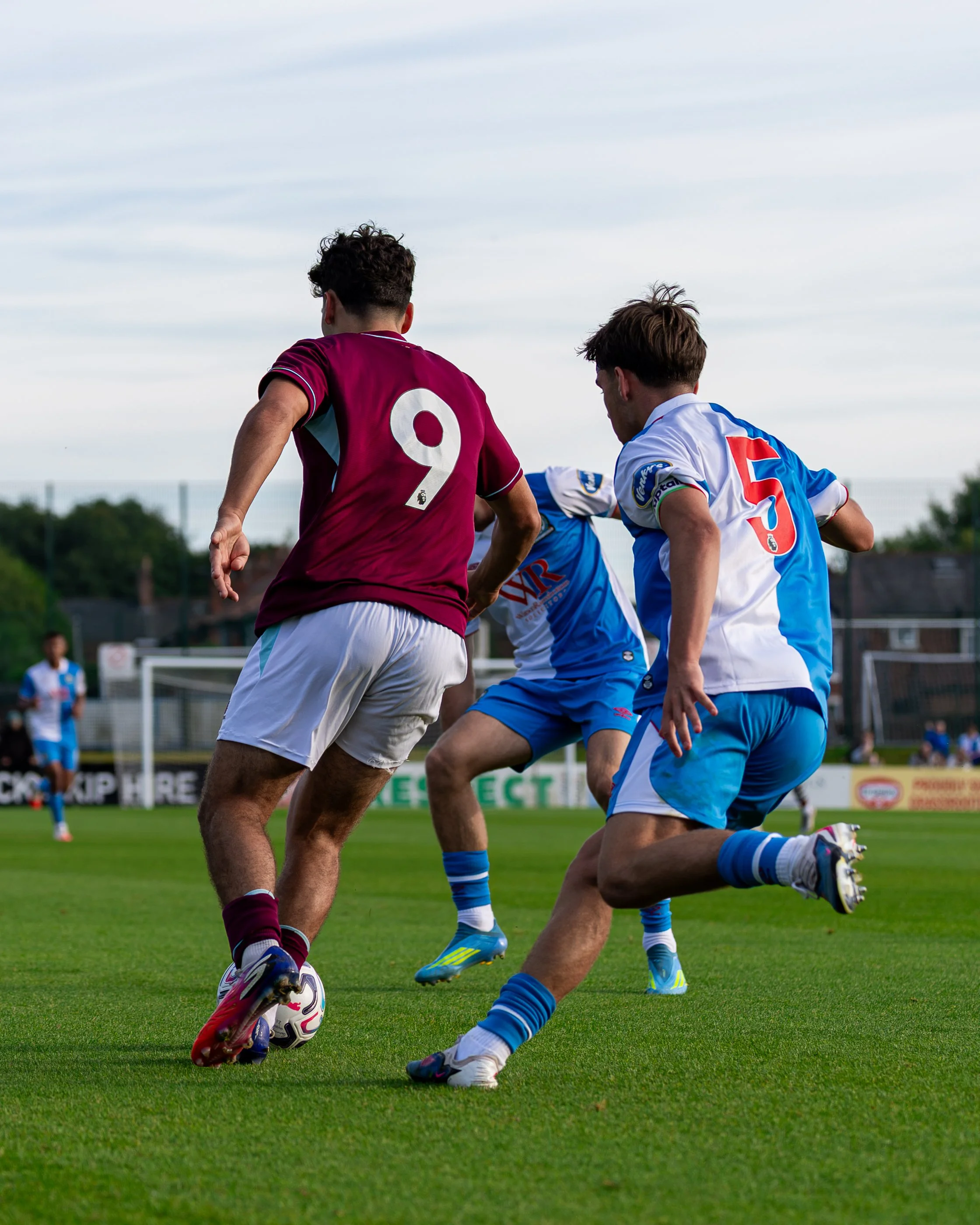 Two soccer players in action on the field, one wearing a maroon jersey with the number 9, and the other in a white and blue jersey with the number 5, competing for the ball.