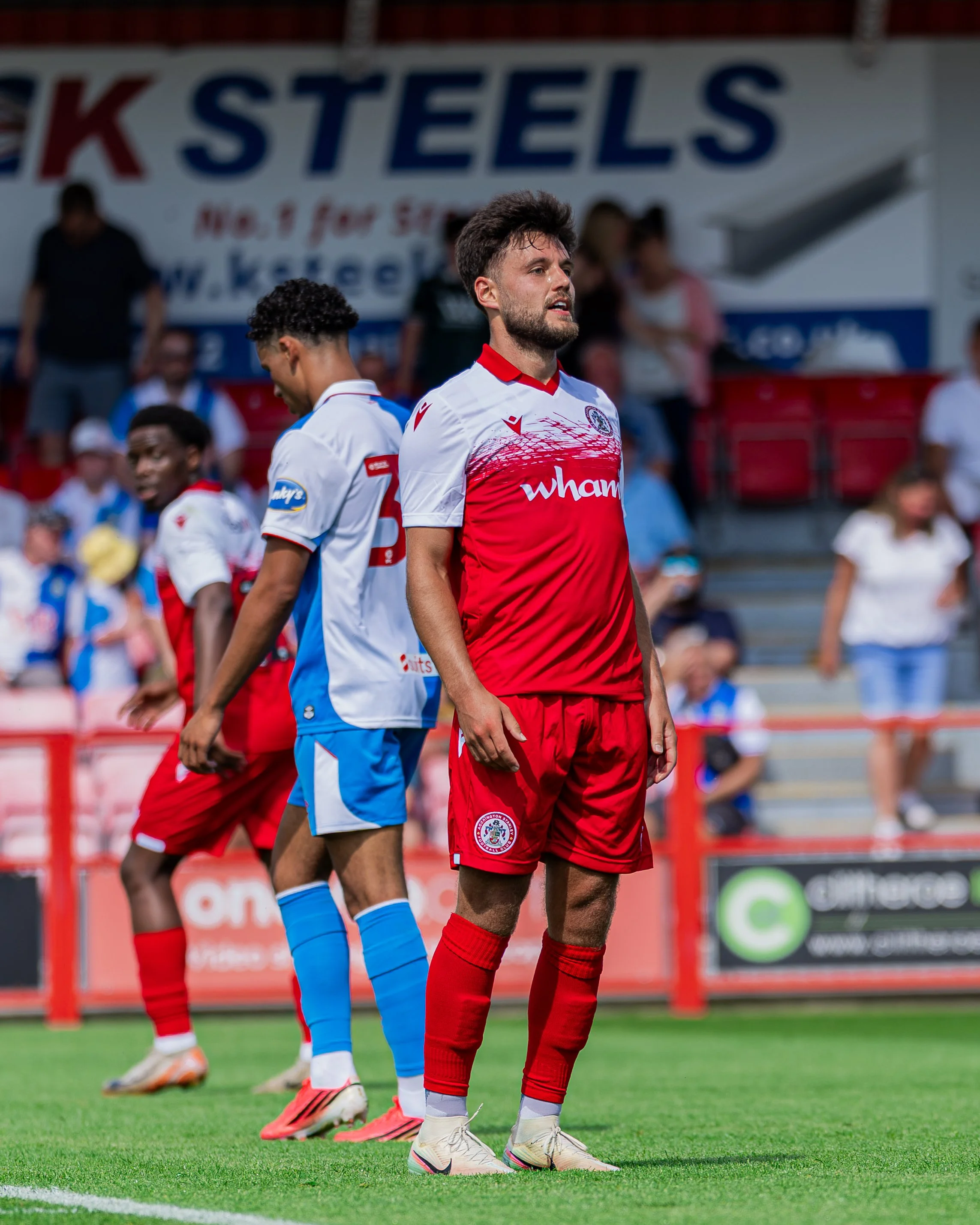 A group of soccer players standing on the field during a match, with one player in a red and white uniform in the foreground, and others in blue and white uniforms in the background.