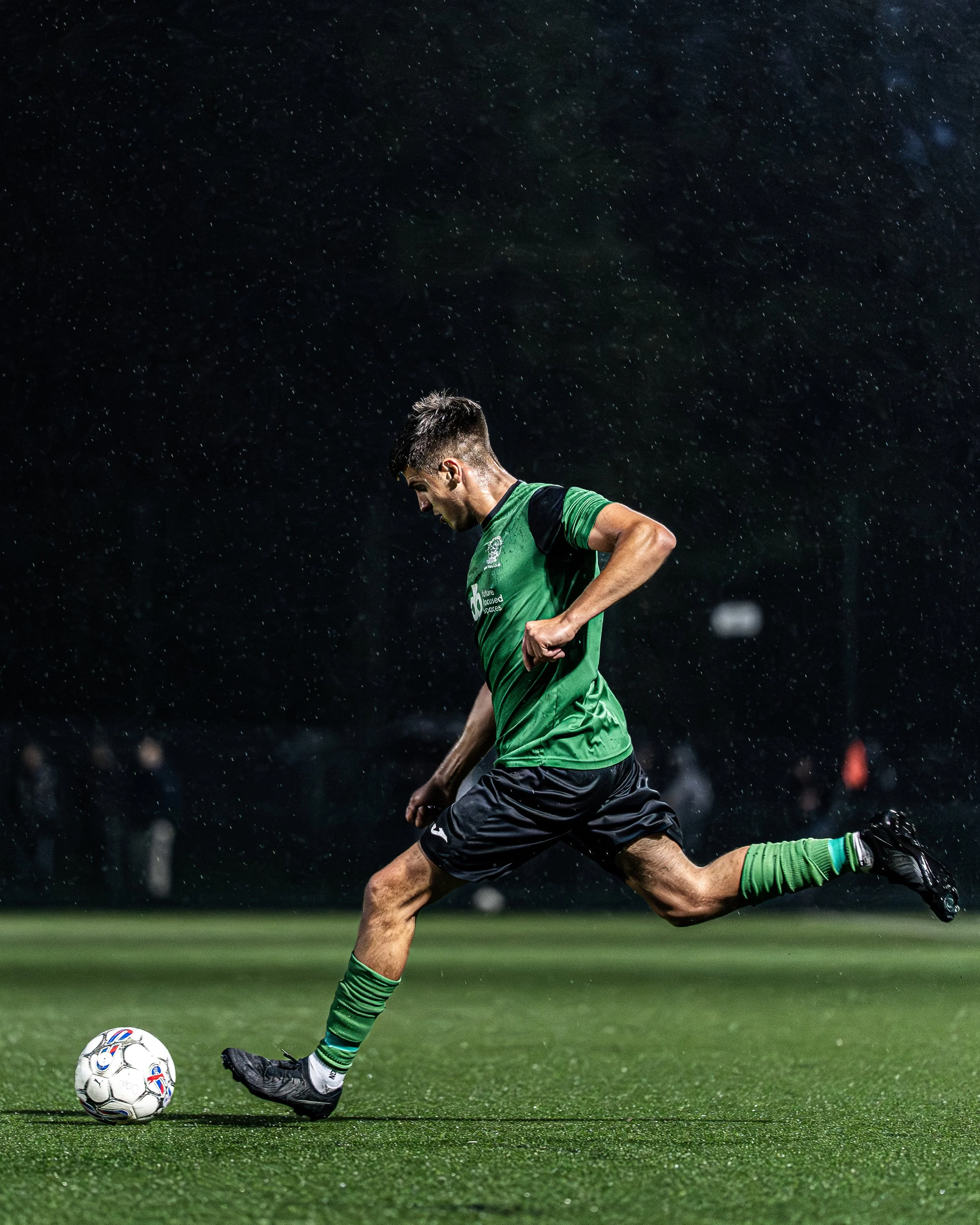 A soccer player in a green uniform kicking a soccer ball on a field during nighttime rain.