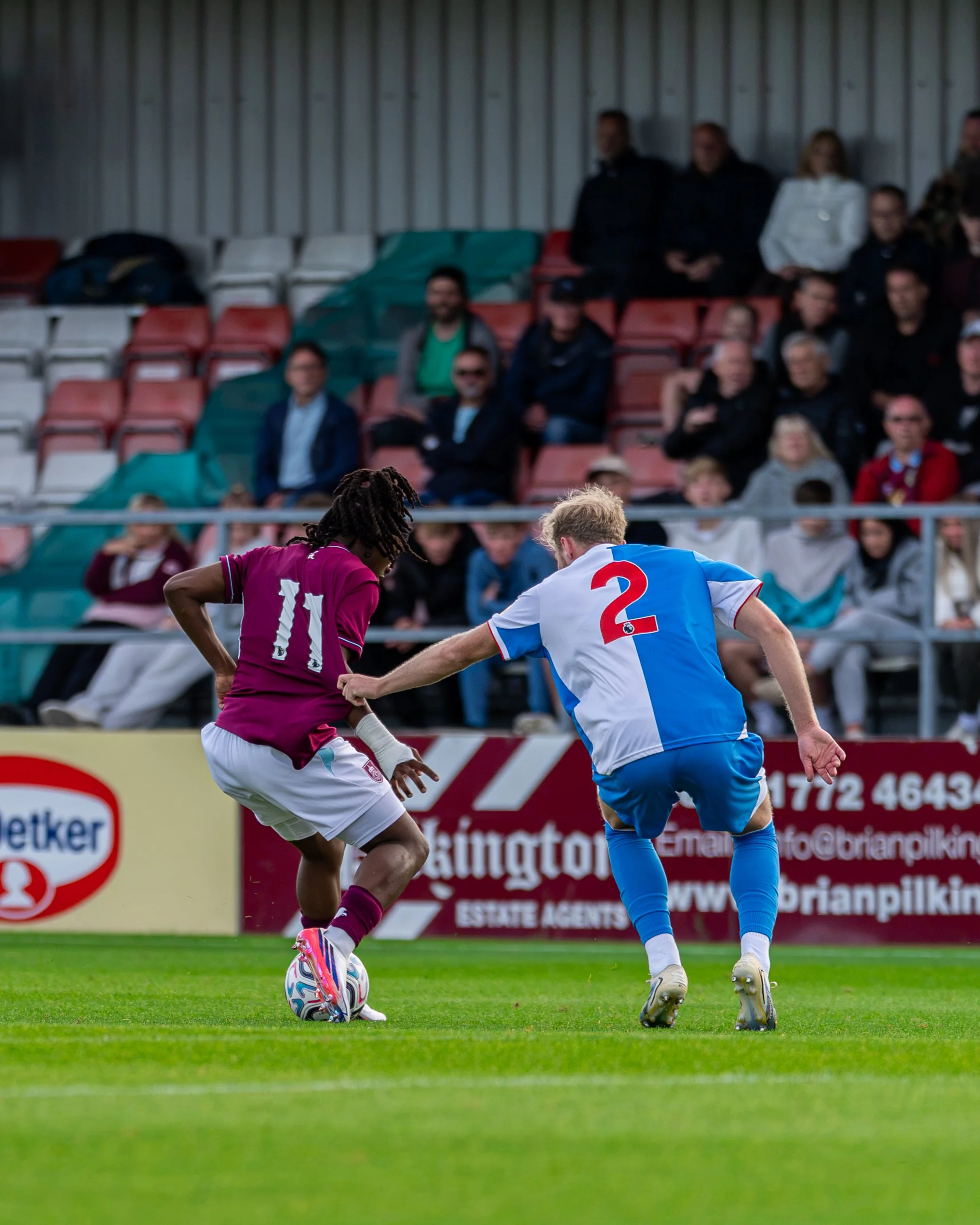 Two soccer players, one in a maroon uniform with the number 11 and the other in a blue and white uniform with the number 2, competing for the ball on a soccer field. Spectators are visible in the background sitting in stadium seats.