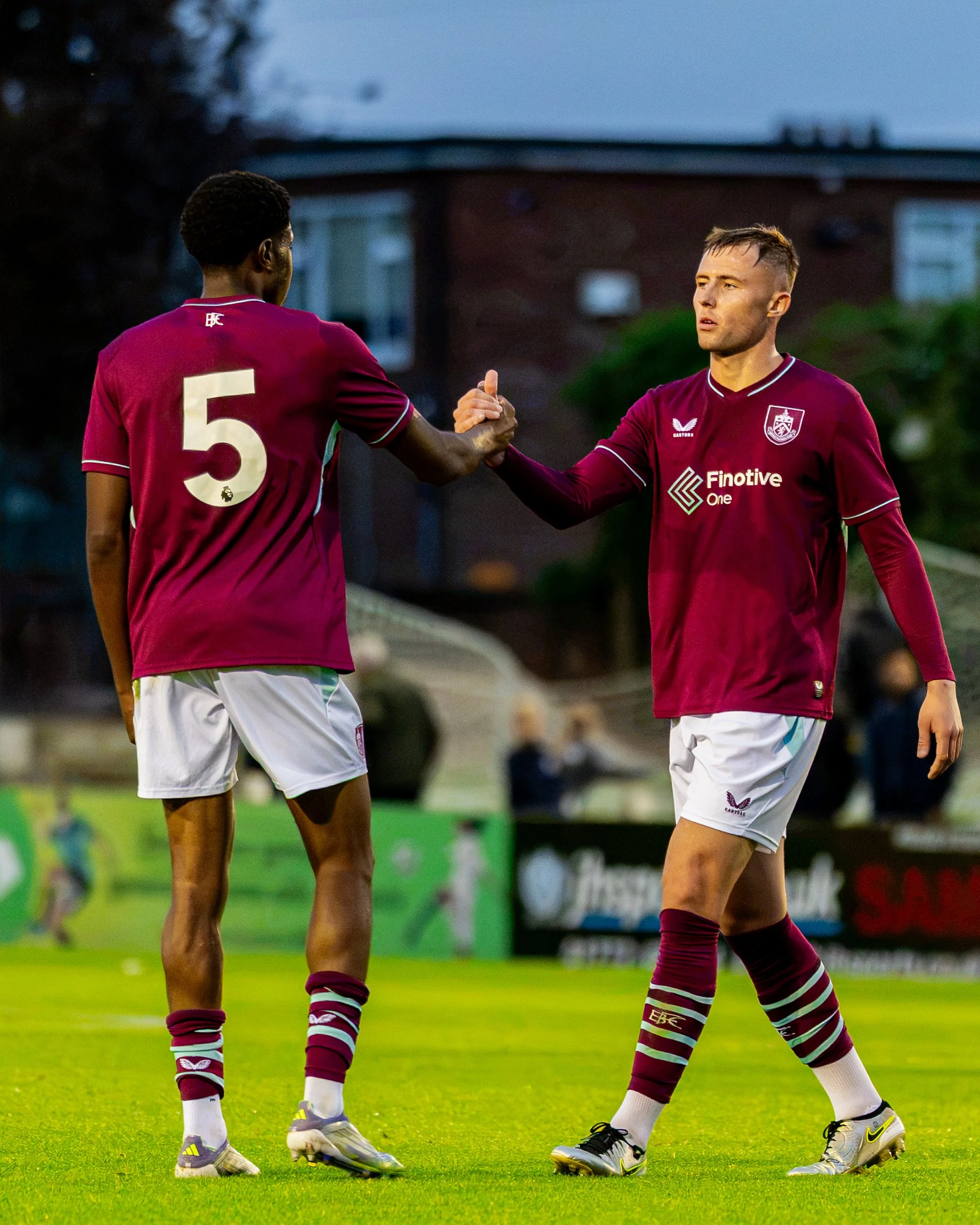 Two soccer players in maroon jerseys shaking hands on a grassy field during daylight, with trees and a building in the background.
