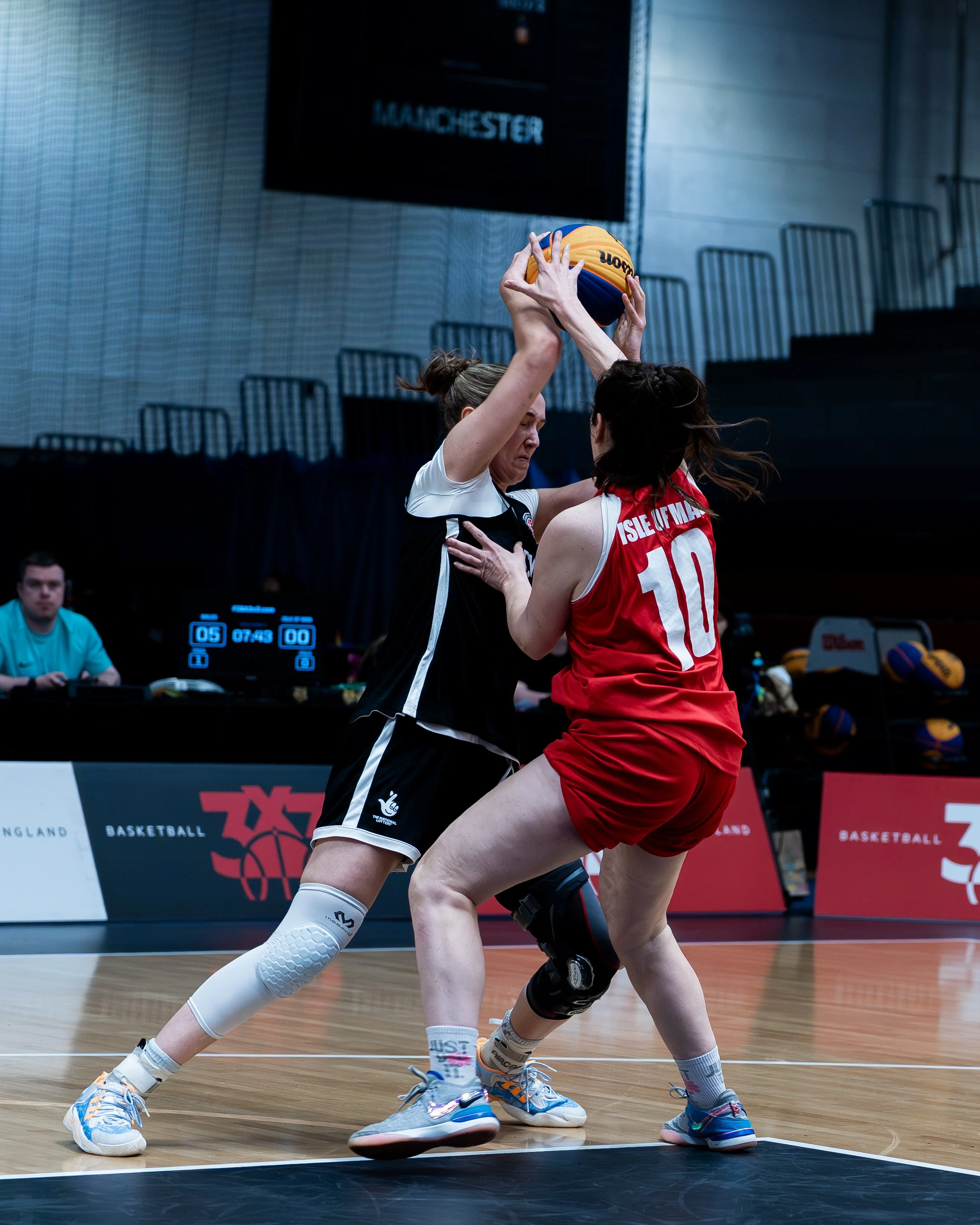Two female basketball players competing for possession of the ball during a game on an indoor court. The player in black is reaching above her head for the ball, while the player in red is trying to block her. A scoreboard is visible in the backgroun