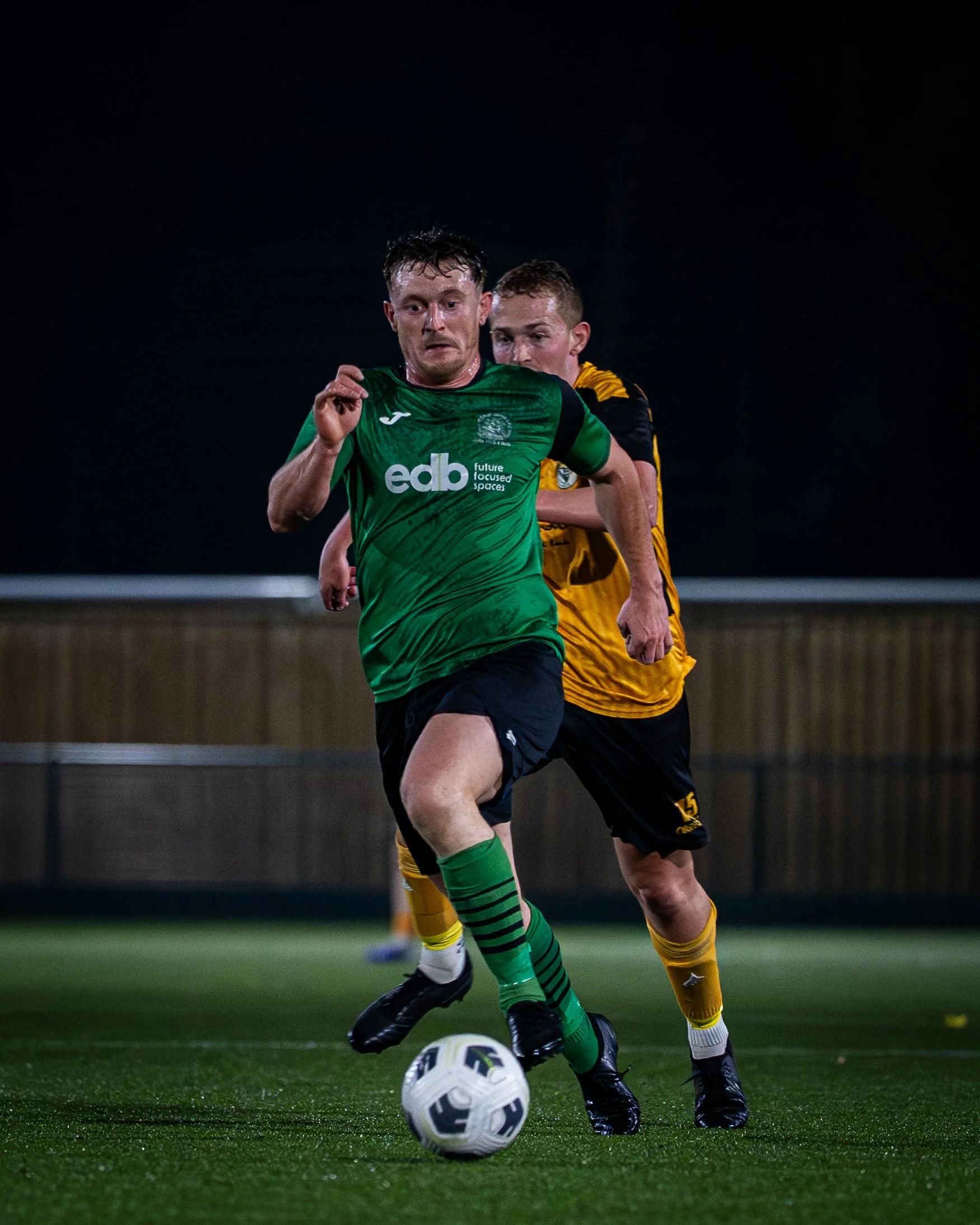 Two soccer players, one in a green uniform and the other in a yellow uniform, compete for the ball during a match on a grassy field at night.
