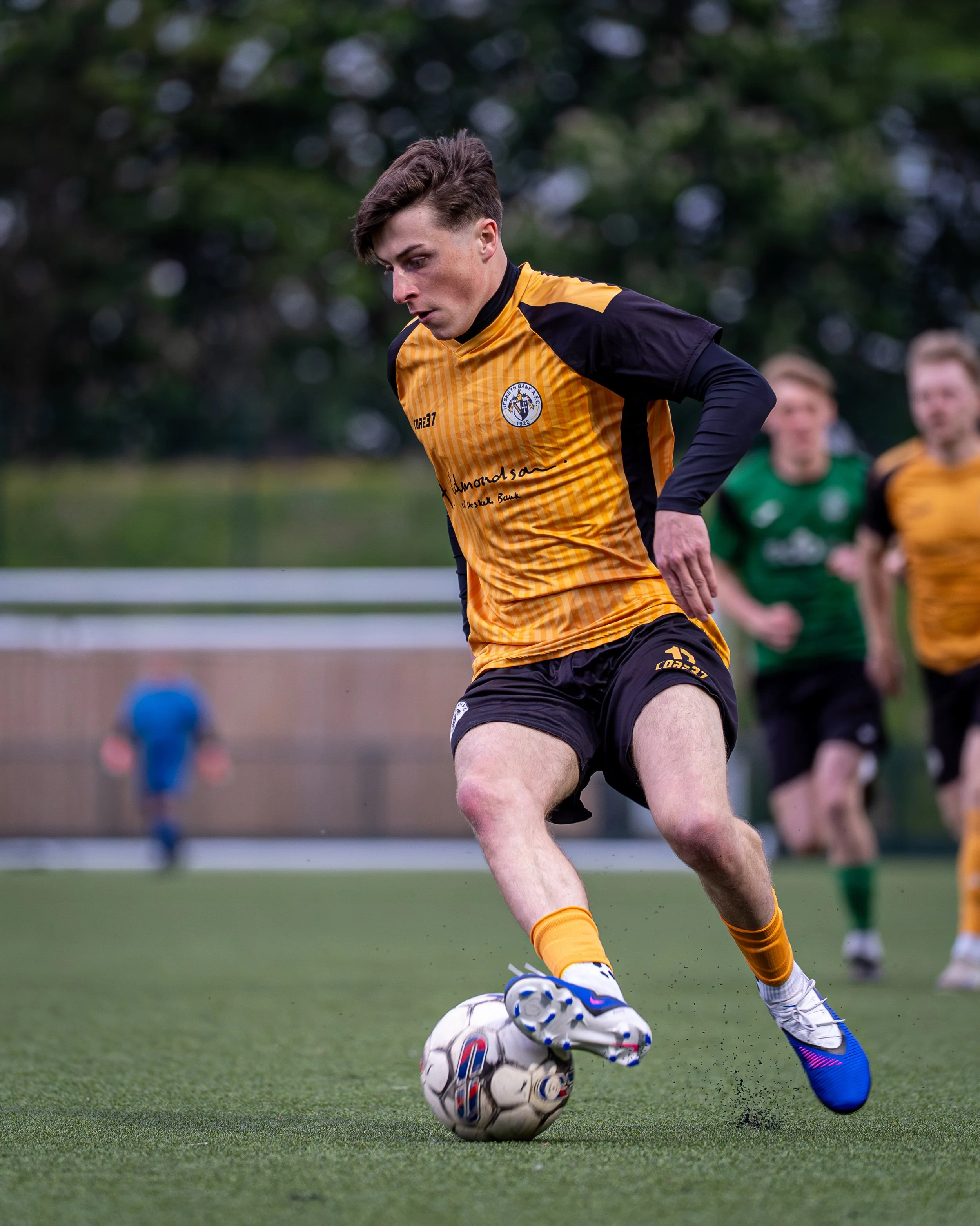 A young male soccer player in yellow and black uniform is kicking a soccer ball on a green field. He is wearing blue and white cleats and yellow socks. Other players are blurred in the background.
