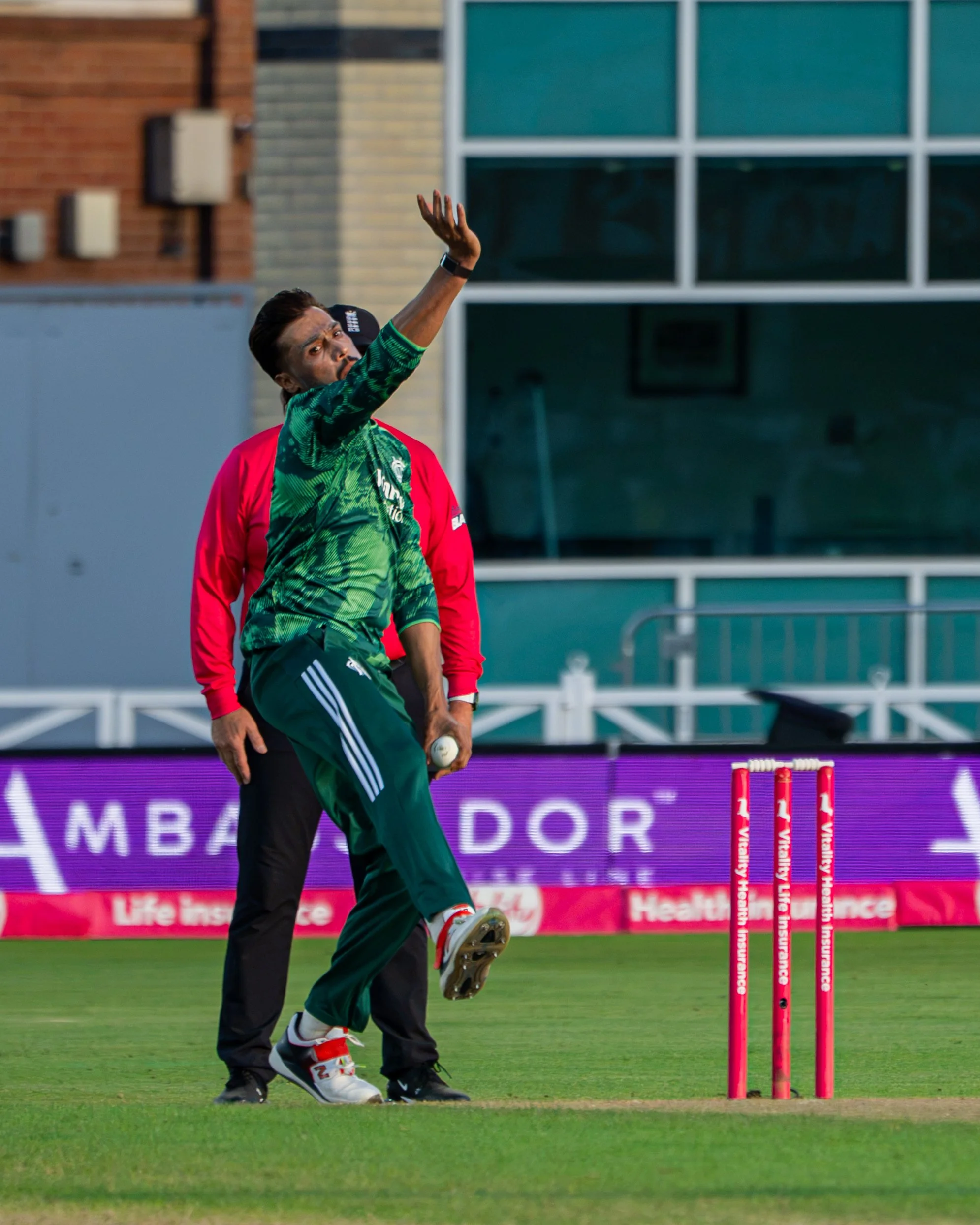 A cricket player in a green and black uniform is bowling a ball on the field, with pink stumps nearby and a person in black in the background.
