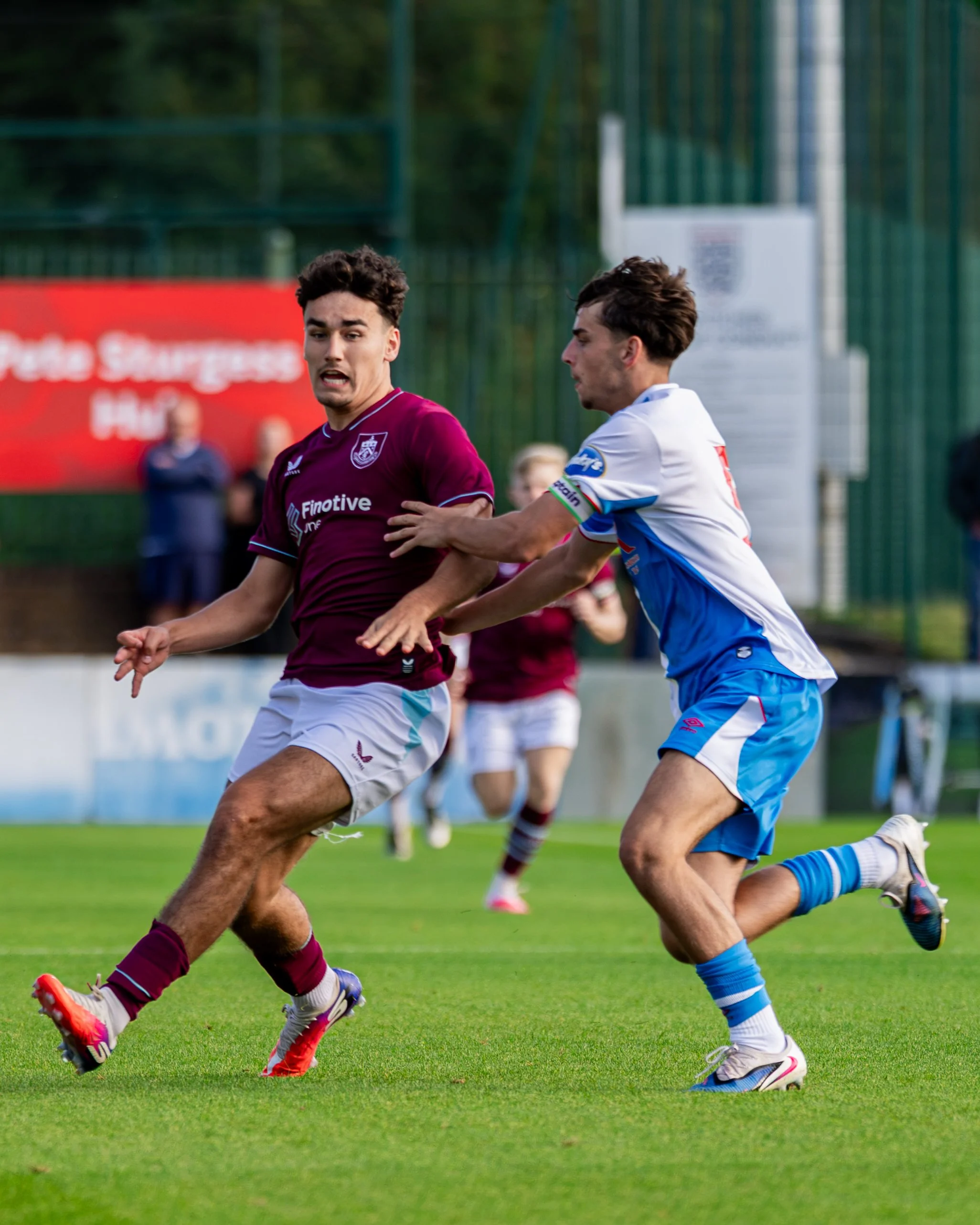 Two soccer players, one wearing a maroon jersey and the other in a white jersey with blue accents, are competing for the ball on a green field, with a red advertising board and some spectators in the background.