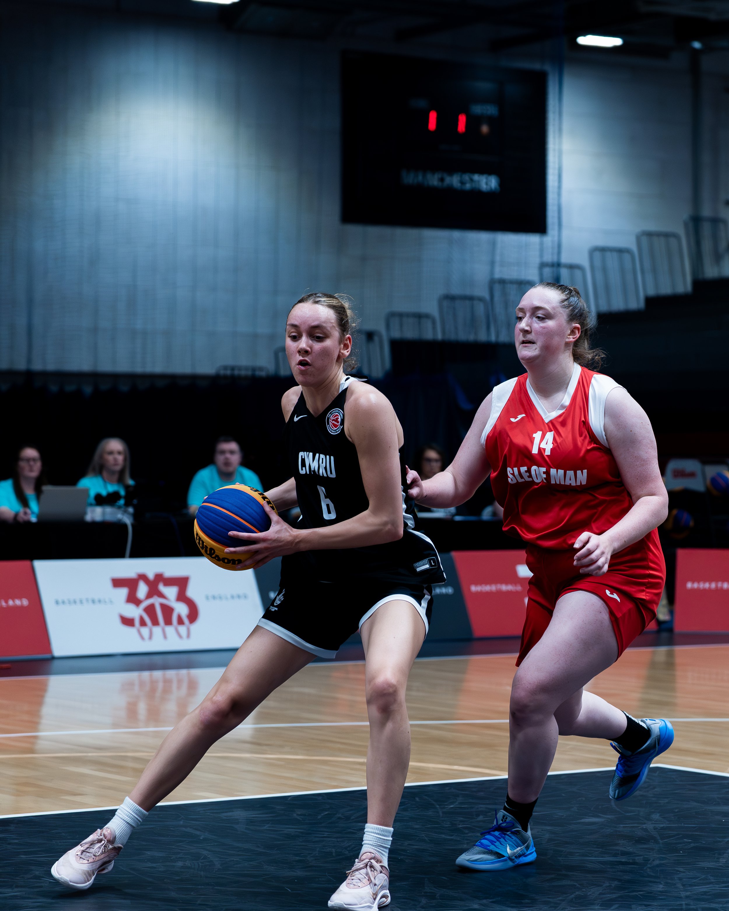 Two female basketball players in a game, one in a black uniform holding a basketball and the other in a red uniform defending, on an indoor court.