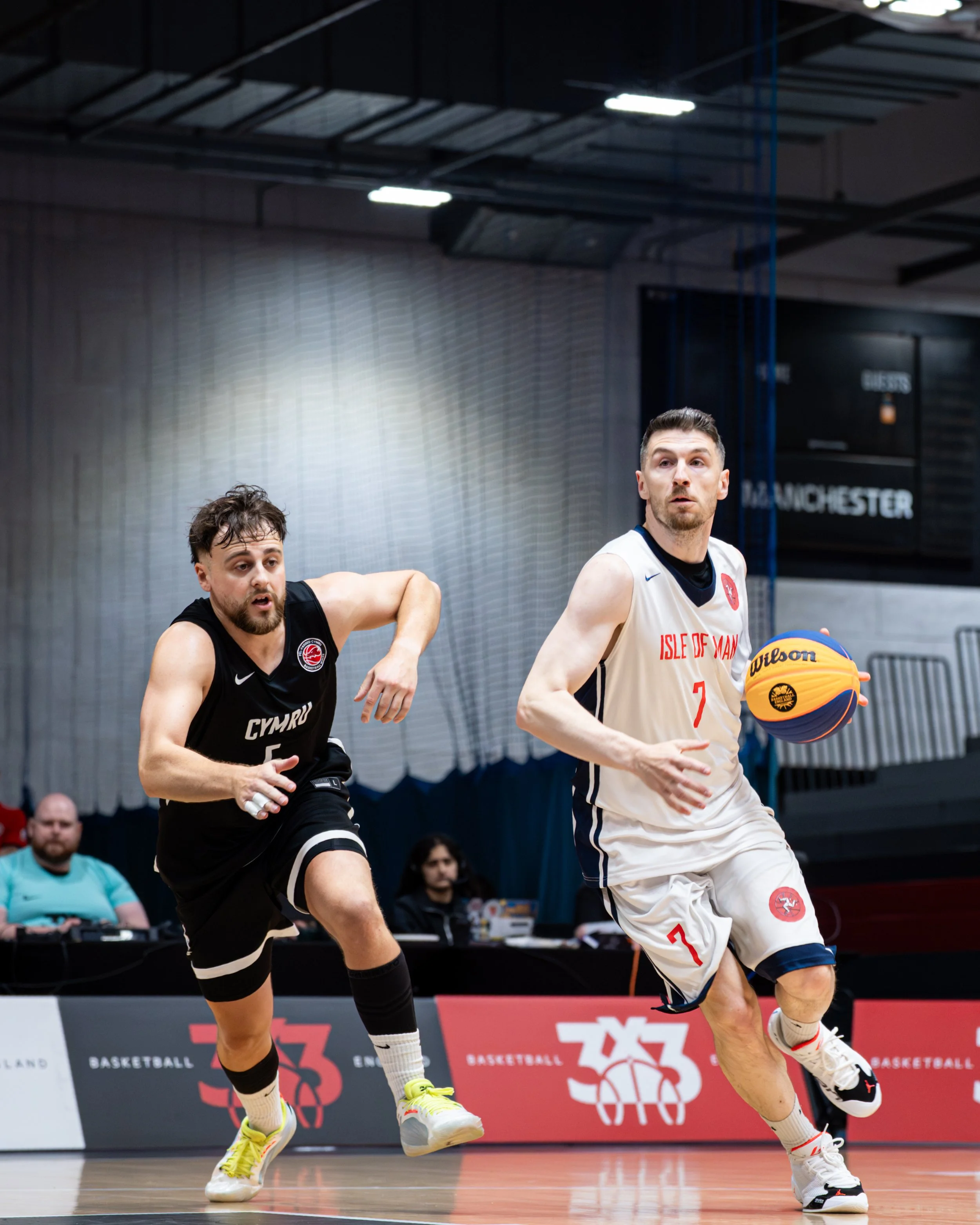 Two basketball players in action during a game. One player, wearing a black uniform with the number 5, is on the left. The other player, wearing a white uniform with the text 'ISLE OF MAN' and the number 7, is on the right, dribbling a Wilson basketb
