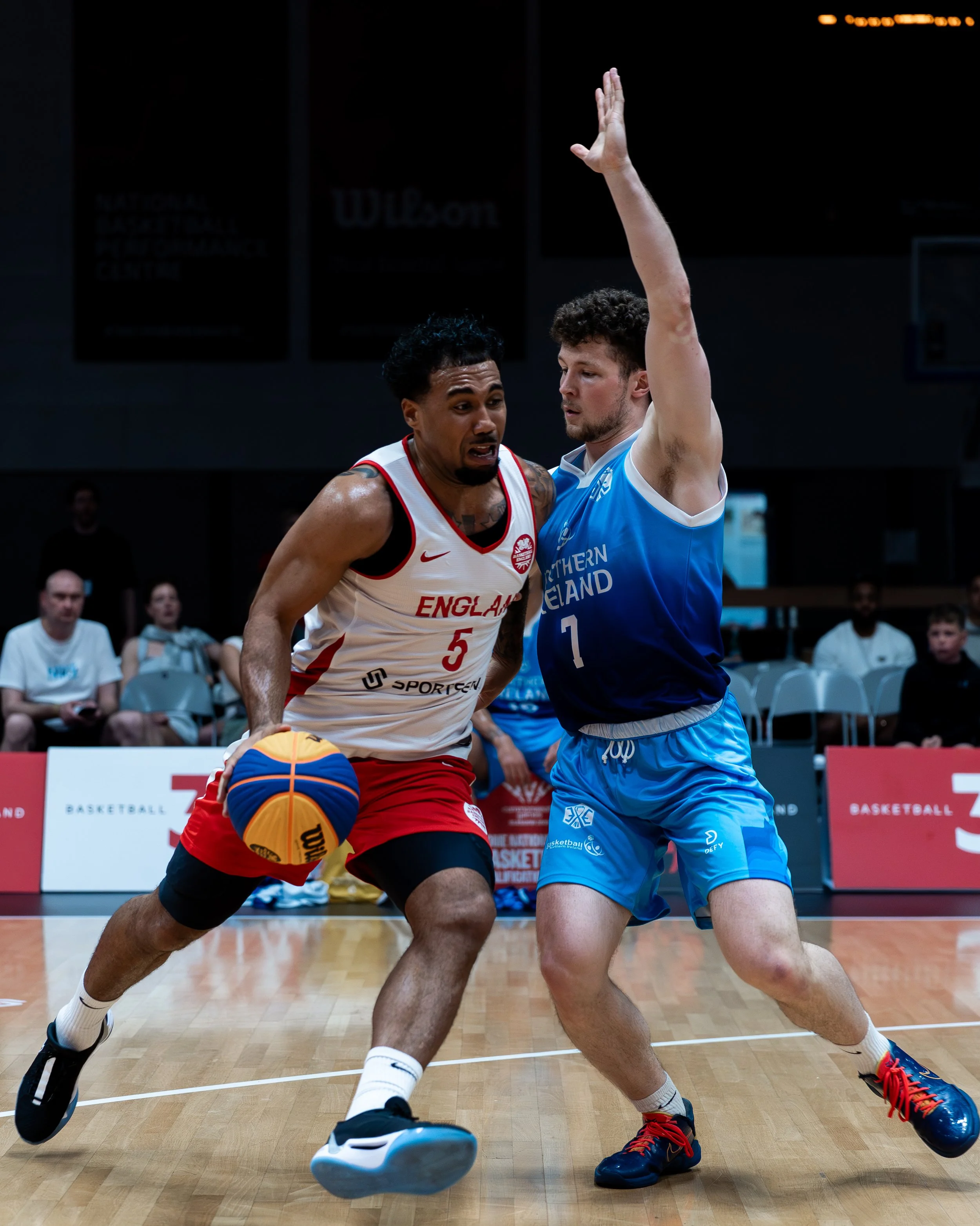 Two male basketball players are fighting for the ball during a game. The player on the left in a white and red uniform is trying to drive the ball while the player on the right in a blue uniform is trying to block or steal it. The background features