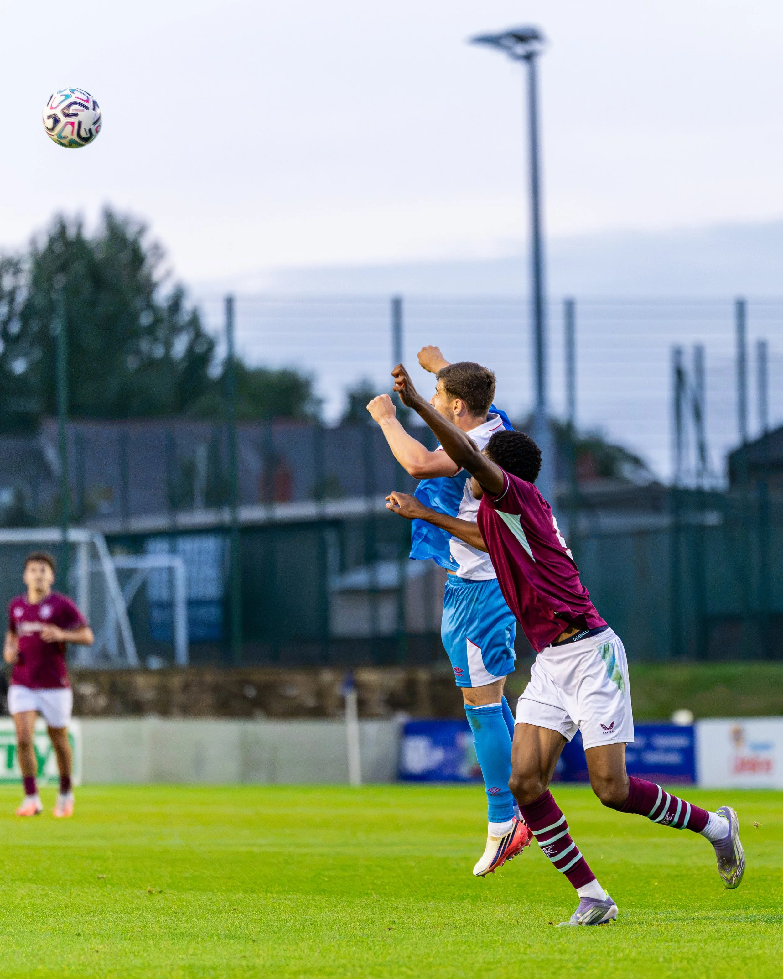 Two soccer players jumping to head the ball during a match on a green field, with one in a blue uniform and the other in a maroon and white uniform. Other players and a goalpost are visible in the background.