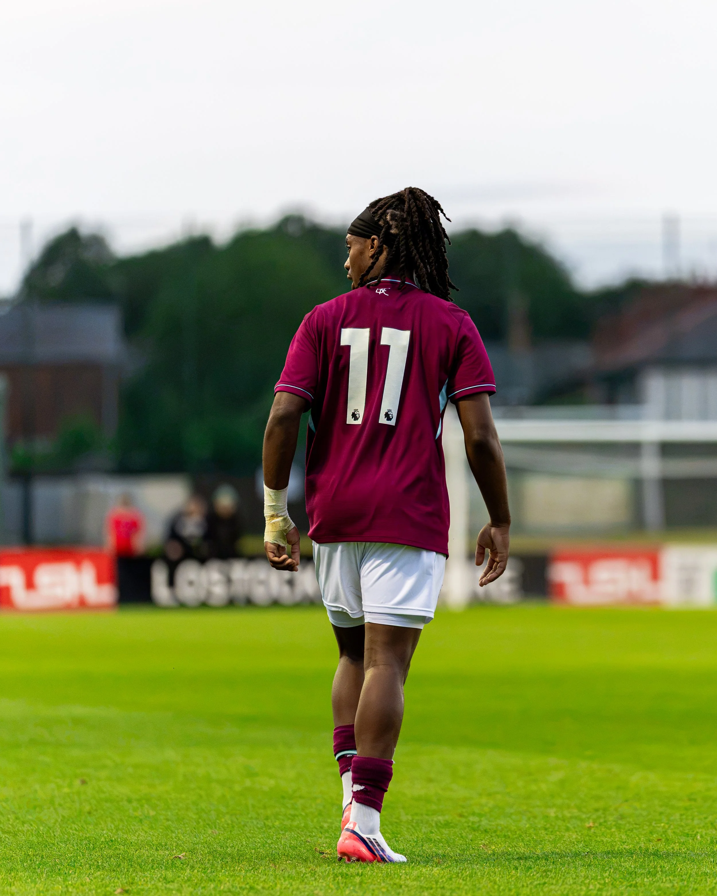 A soccer player wearing a maroon jersey with the number 11 on the back, white shorts, maroon socks with white stripes, and orange cleats standing on a grassy field during a match.