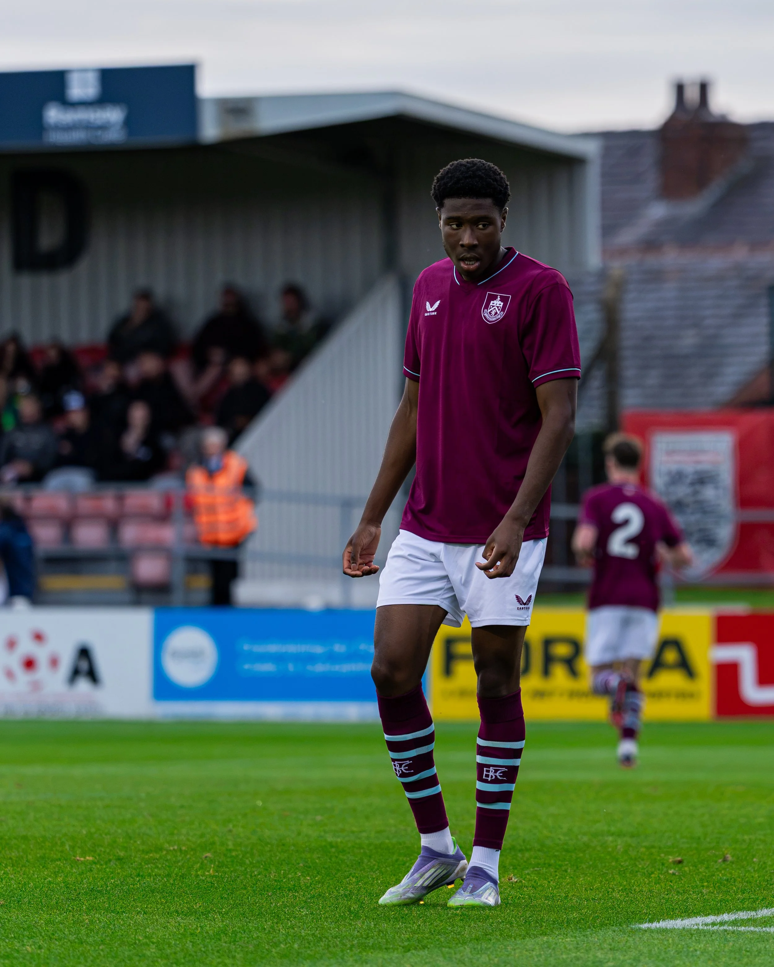 A young male soccer player wearing a maroon jersey and white shorts standing on a green field during a game. There are spectators and advertisements in the background.