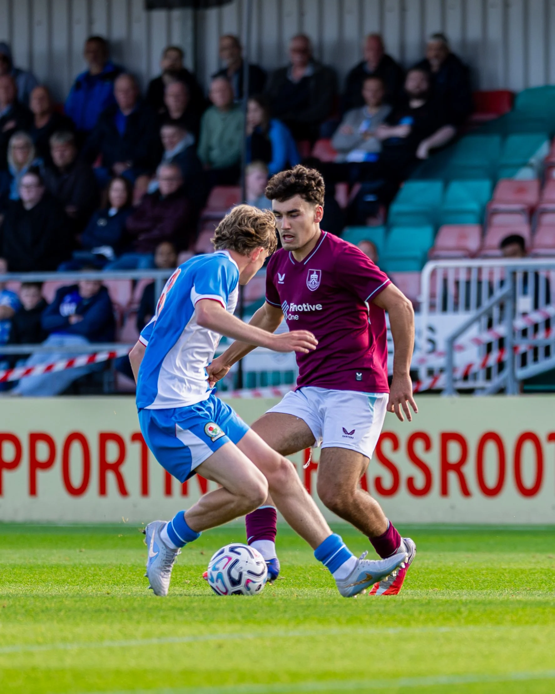 Two soccer players competing for the ball on a field, with a blurred crowd watching in the background.