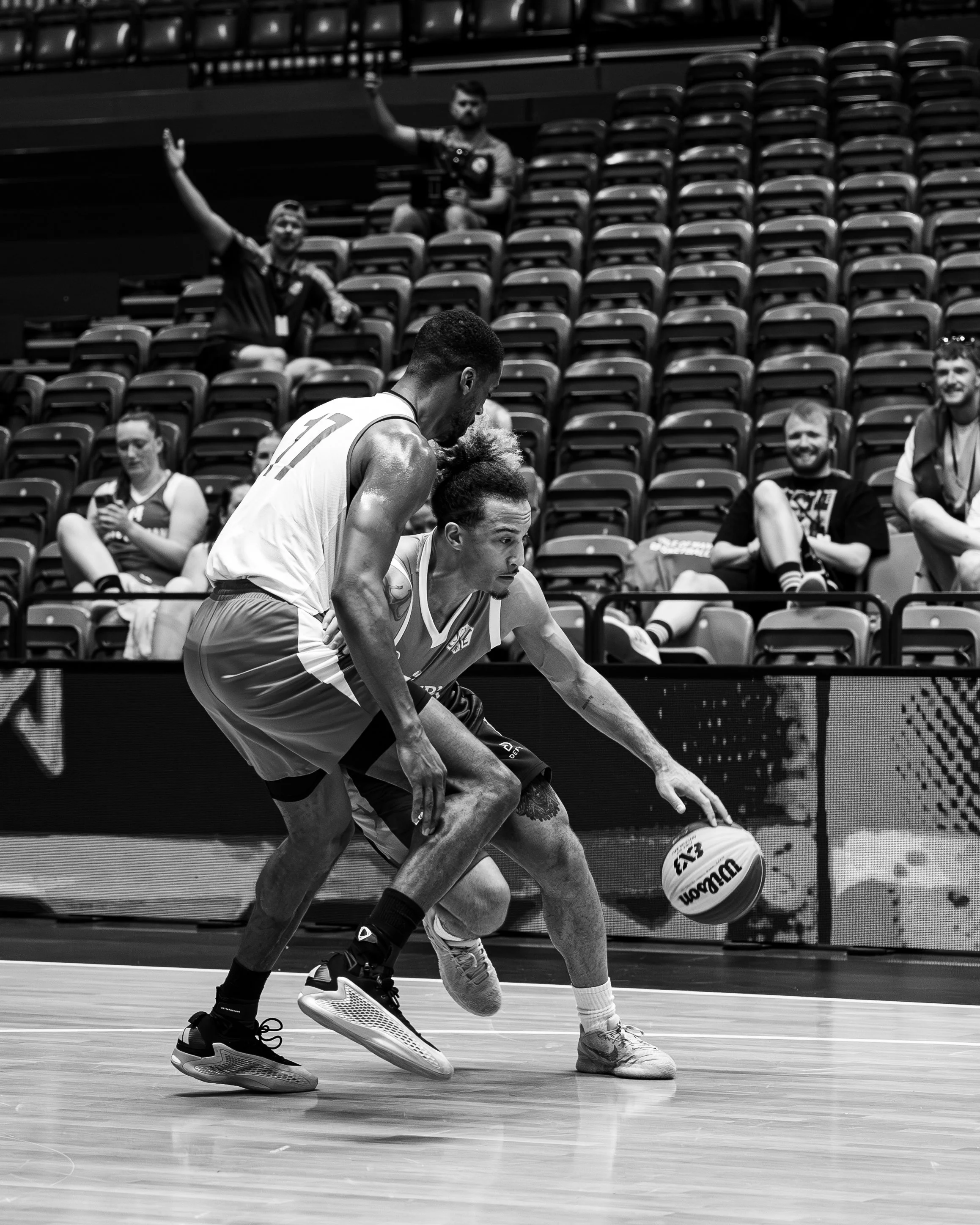 Two basketball players are competing for control of the ball on a basketball court, with spectators watching in the background.