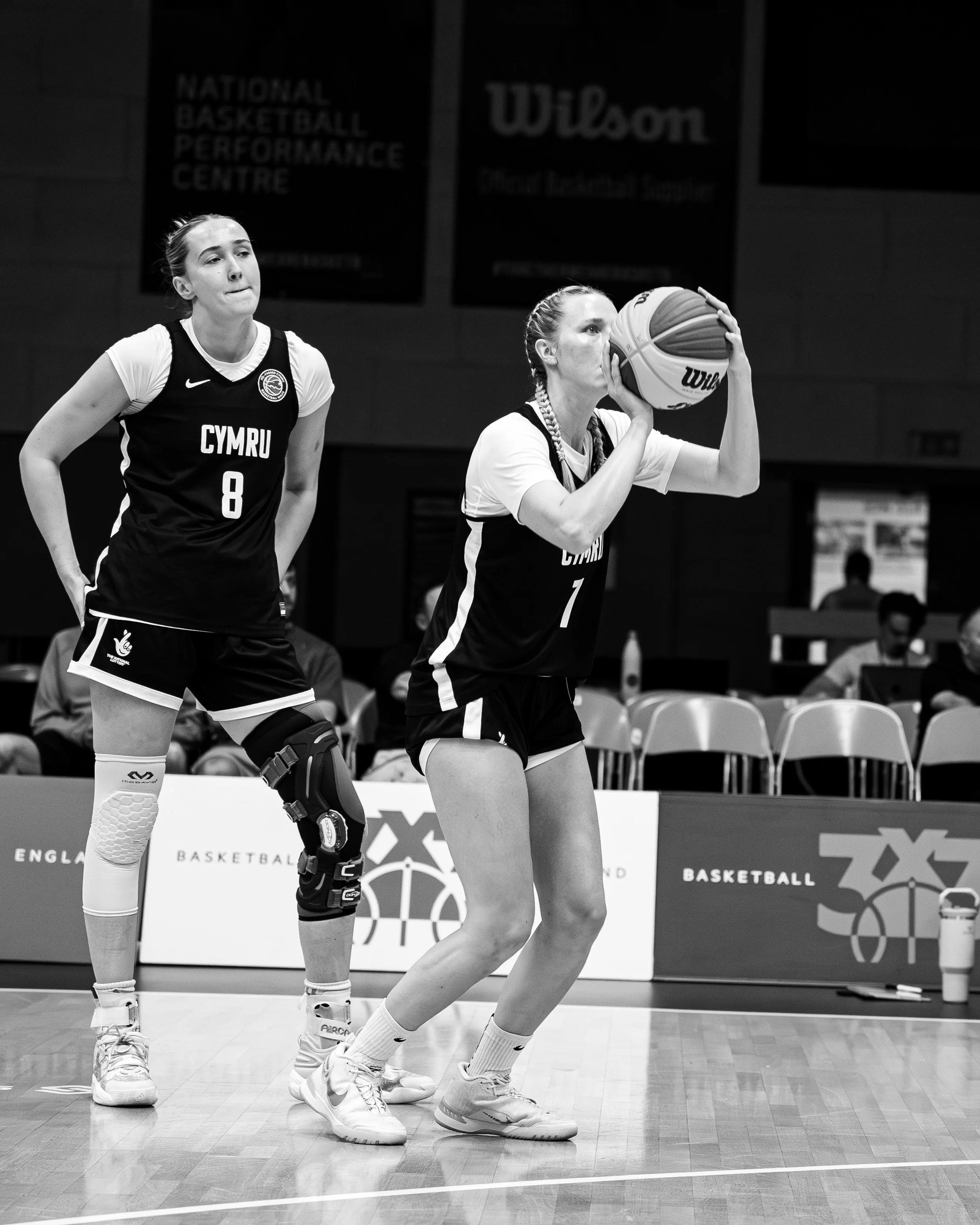 Two female basketball players in action during a game, one preparing to shoot the ball and the other standing nearby, on an indoor court with signage in the background.