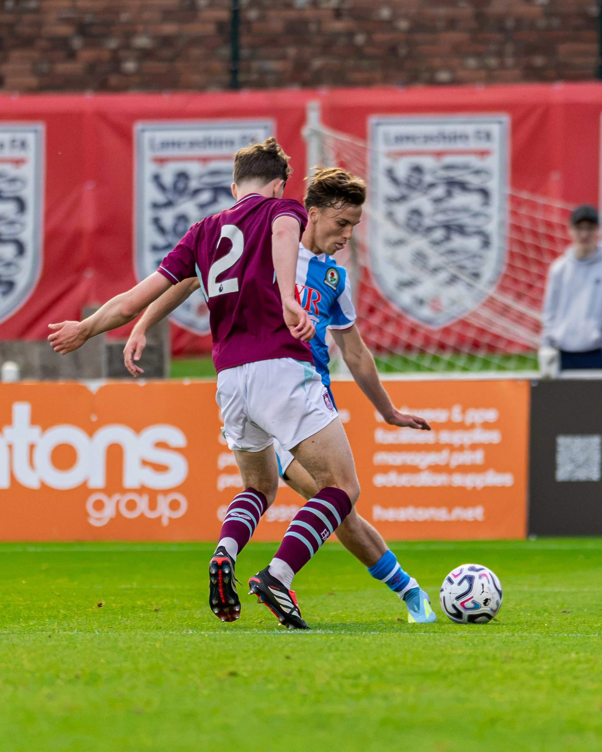 A soccer player wearing a blue jersey and white shorts is kicking a soccer ball on a grass field, with another player in a maroon jersey nearby. There are banners and a brick wall in the background.
