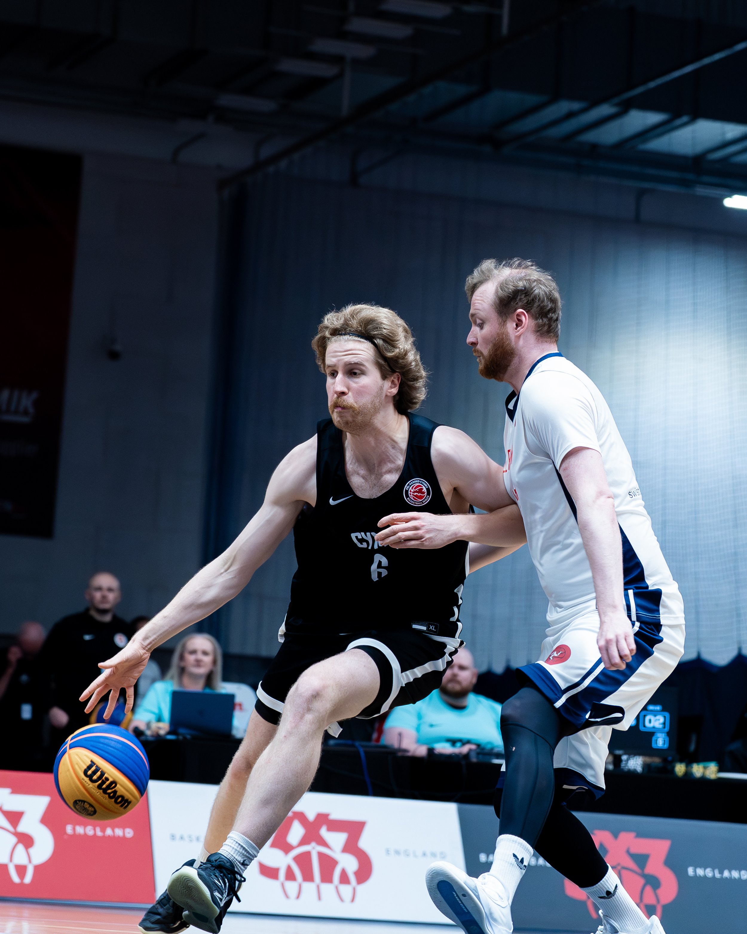 Two male basketball players in action during a game. The player on the left is wearing a black uniform and is reaching for the ball, while the player on the right, in a white uniform, is closely guarding him. The scene takes place indoors with an aud