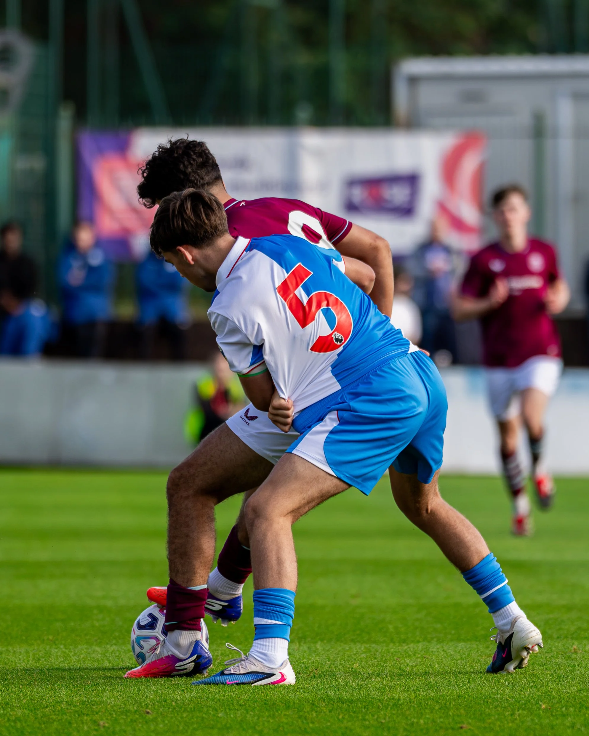 Two male soccer players competing for the ball on the field, one wearing a maroon and white uniform and the other a white and blue uniform, with spectators and banners in the background.