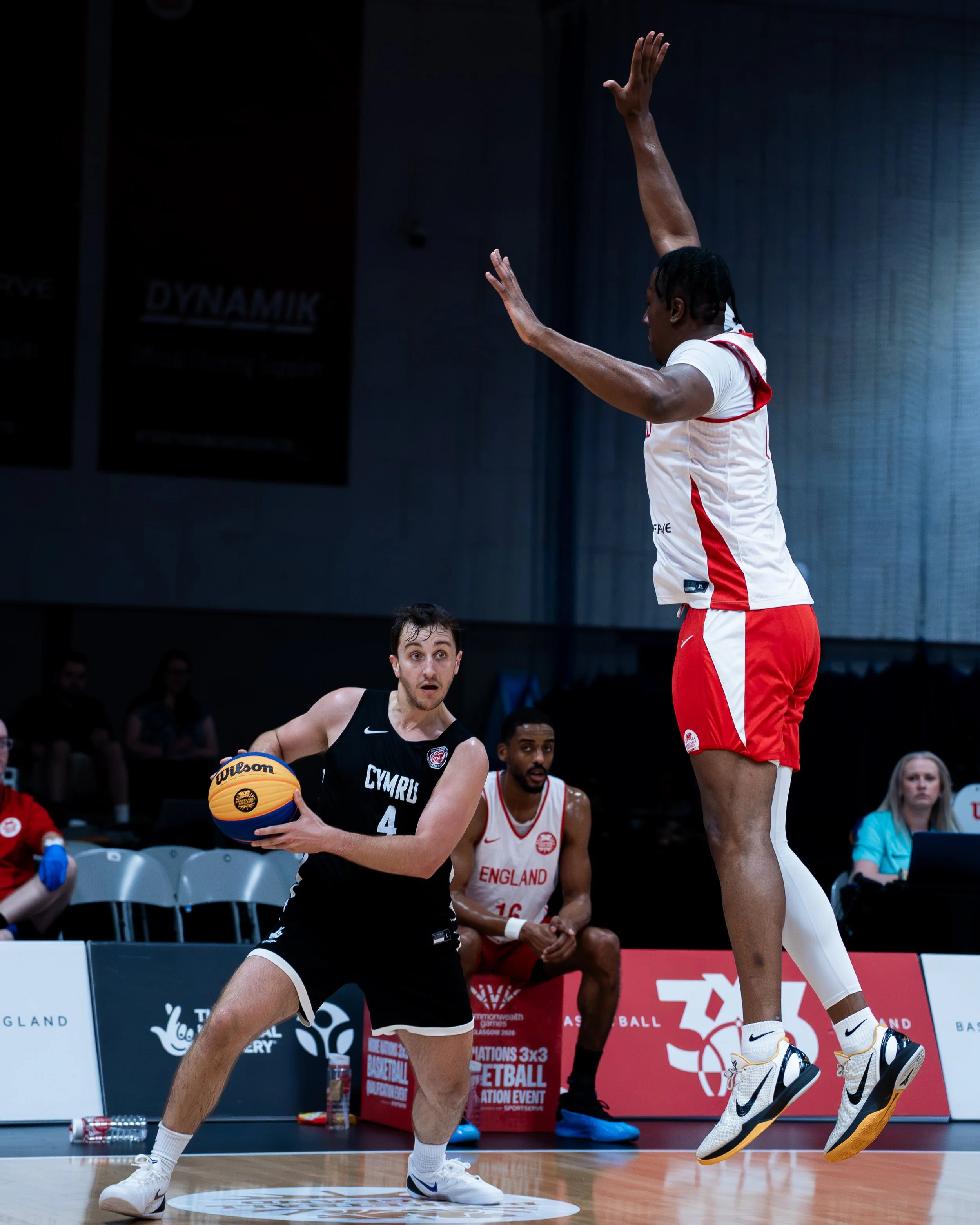 A basketball game featuring players from the England and Wales teams. An England player in a black uniform with the number 4 holds the ball, while a Wales player in a white and red uniform jumps to block or defend. The scene takes place on an indoor 